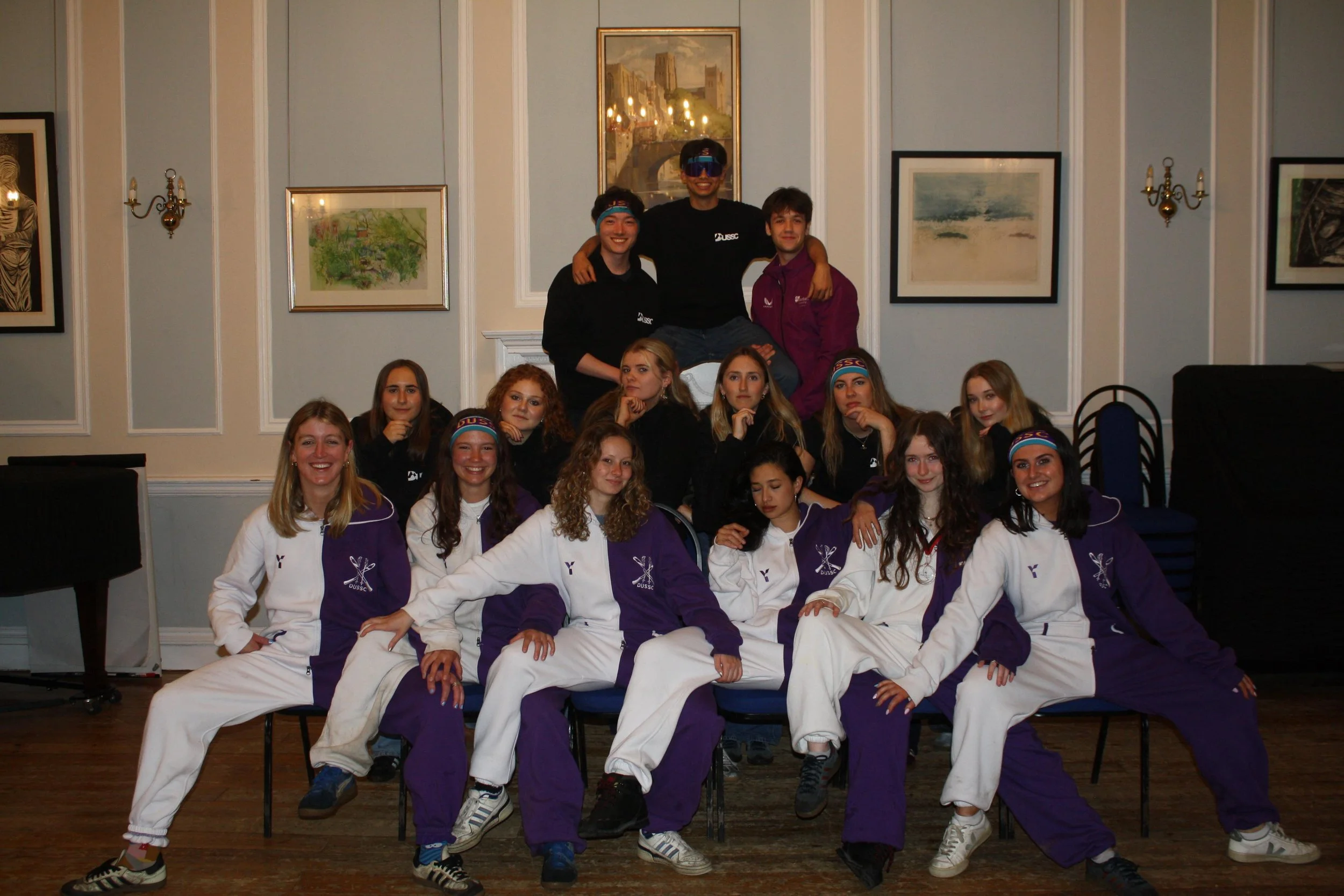 A group of young people posing for a photo in a room with artwork on the walls. Some are sitting and others are standing, with two individuals in the middle standing on chairs. Everyone is smiling or looking at the camera, wearing casual sportswear, some with matching jackets and headbands.