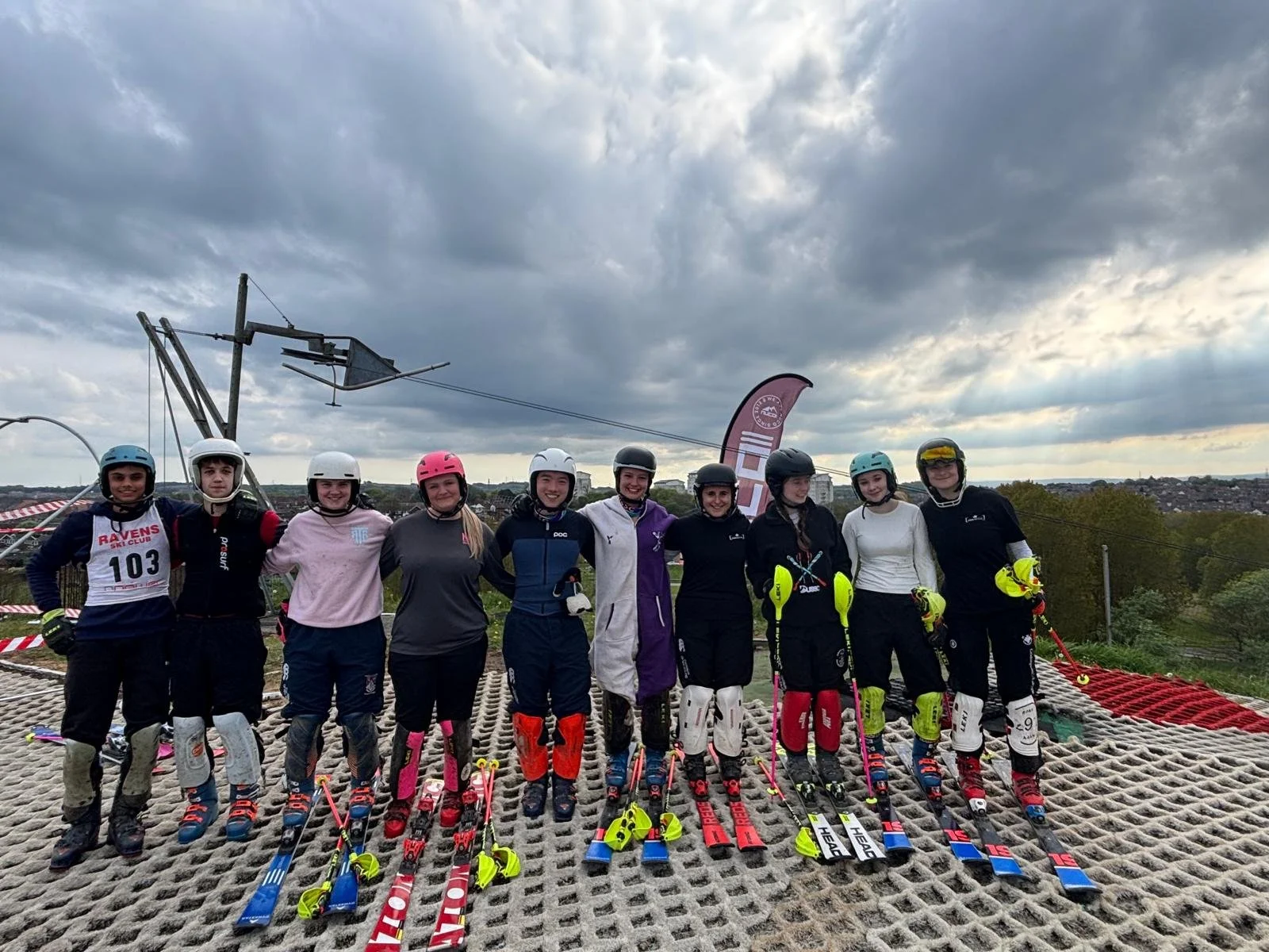 Group of women skiers in ski gear standing together on a ski slope, with skis on the ground in front of them, under a cloudy sky.