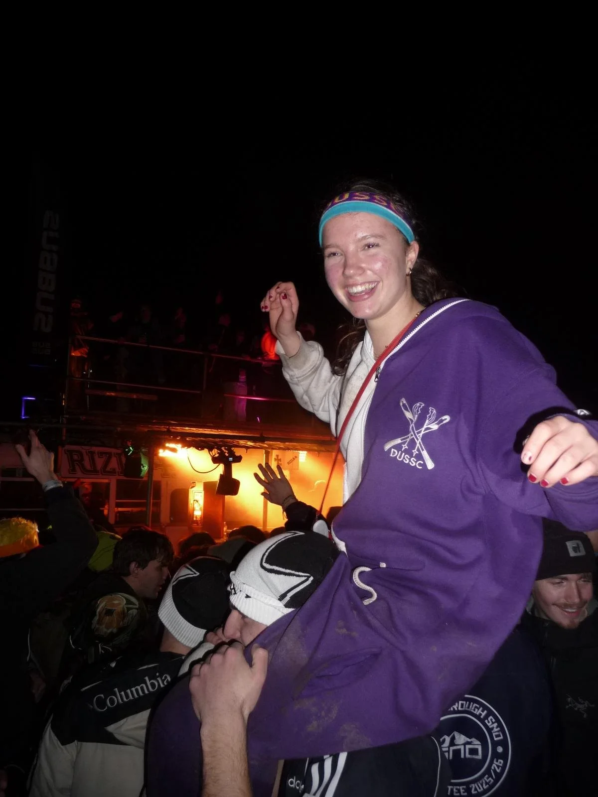 A young woman with braces, wearing a purple hoodie and a blue headband, sitting on someone's shoulders at a nighttime outdoor event, smiling and dancing with people in the background near a stage with bright orange lighting.
