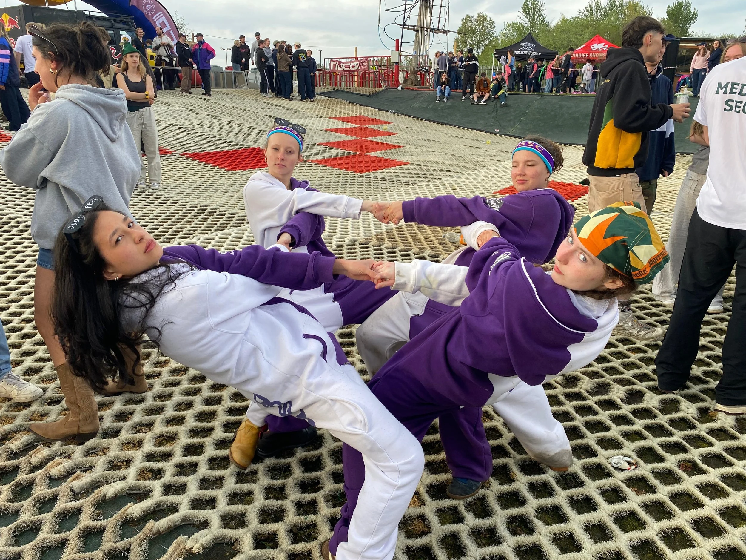 Six young women wearing purple and white outfits are lying on their backs on a netted surface, holding hands, at an outdoor event. People are standing and sitting nearby, with tents, banners, and rides in the background.