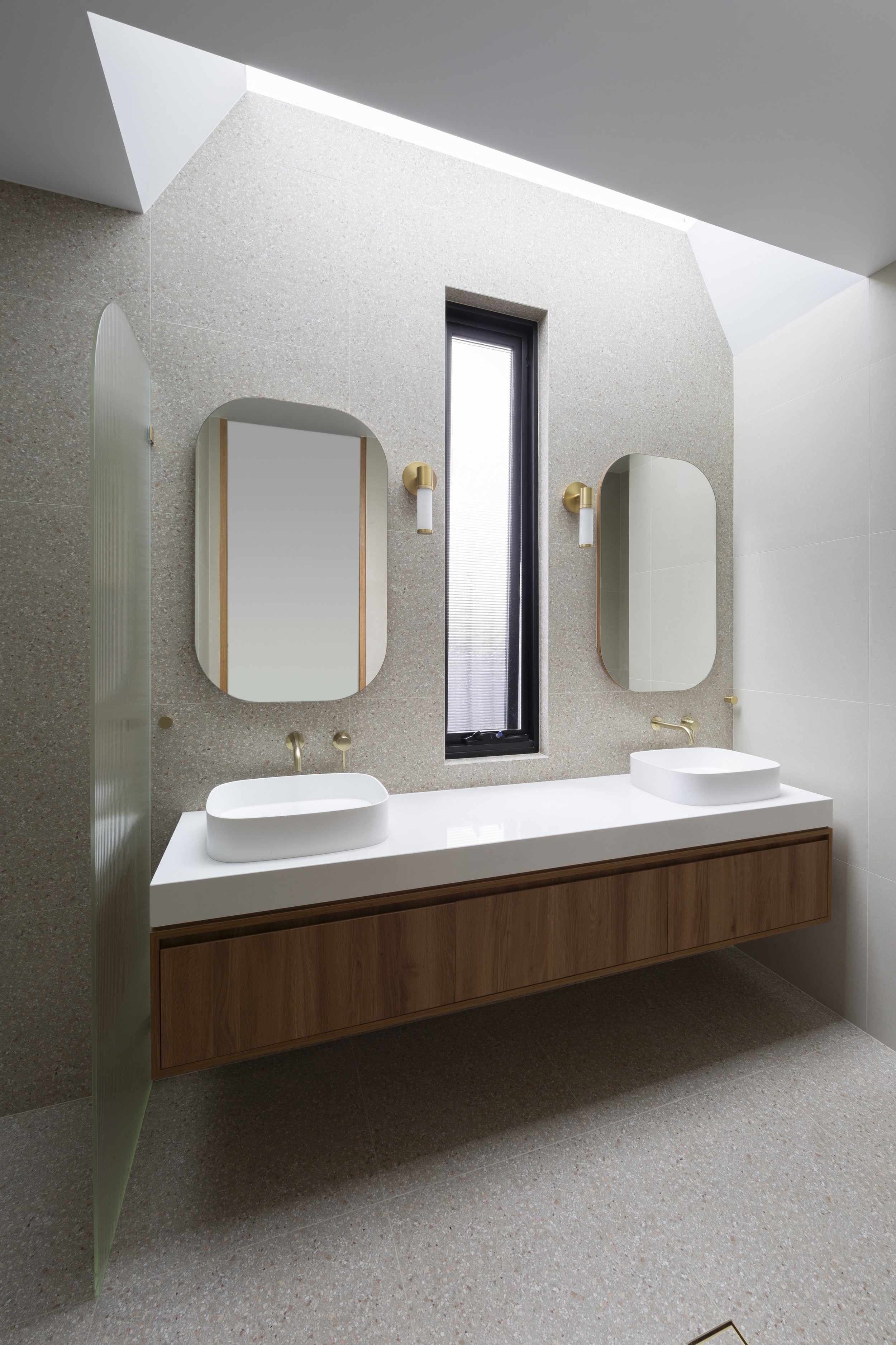 Modern bathroom with double vessel sinks, wooden vanity, rectangular mirrors, black window, and gold fixtures on a terrazzo wall.
