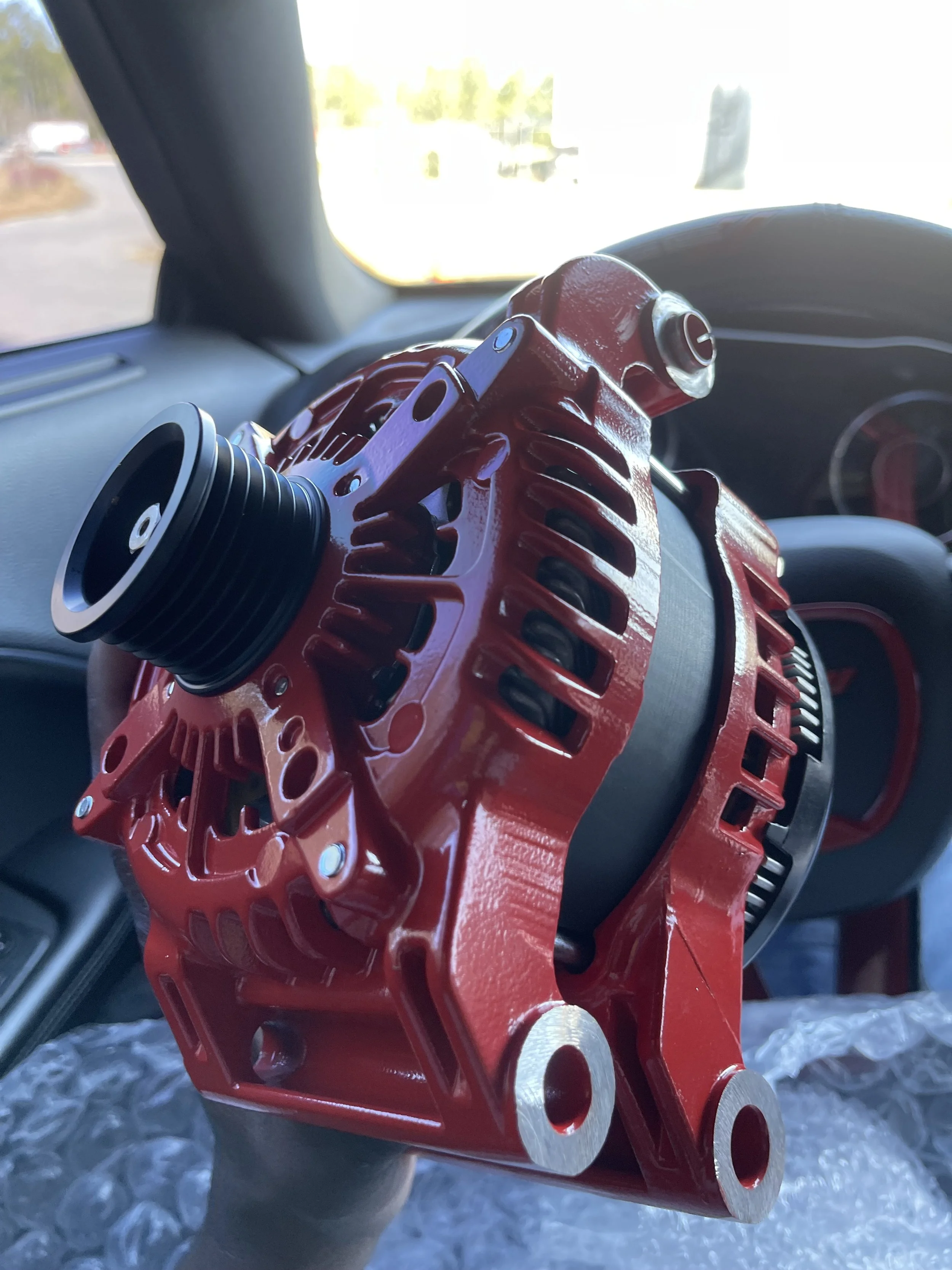 A red mechanical part, possibly an engine component, being held inside a car with a blurred outdoor scene in the background.