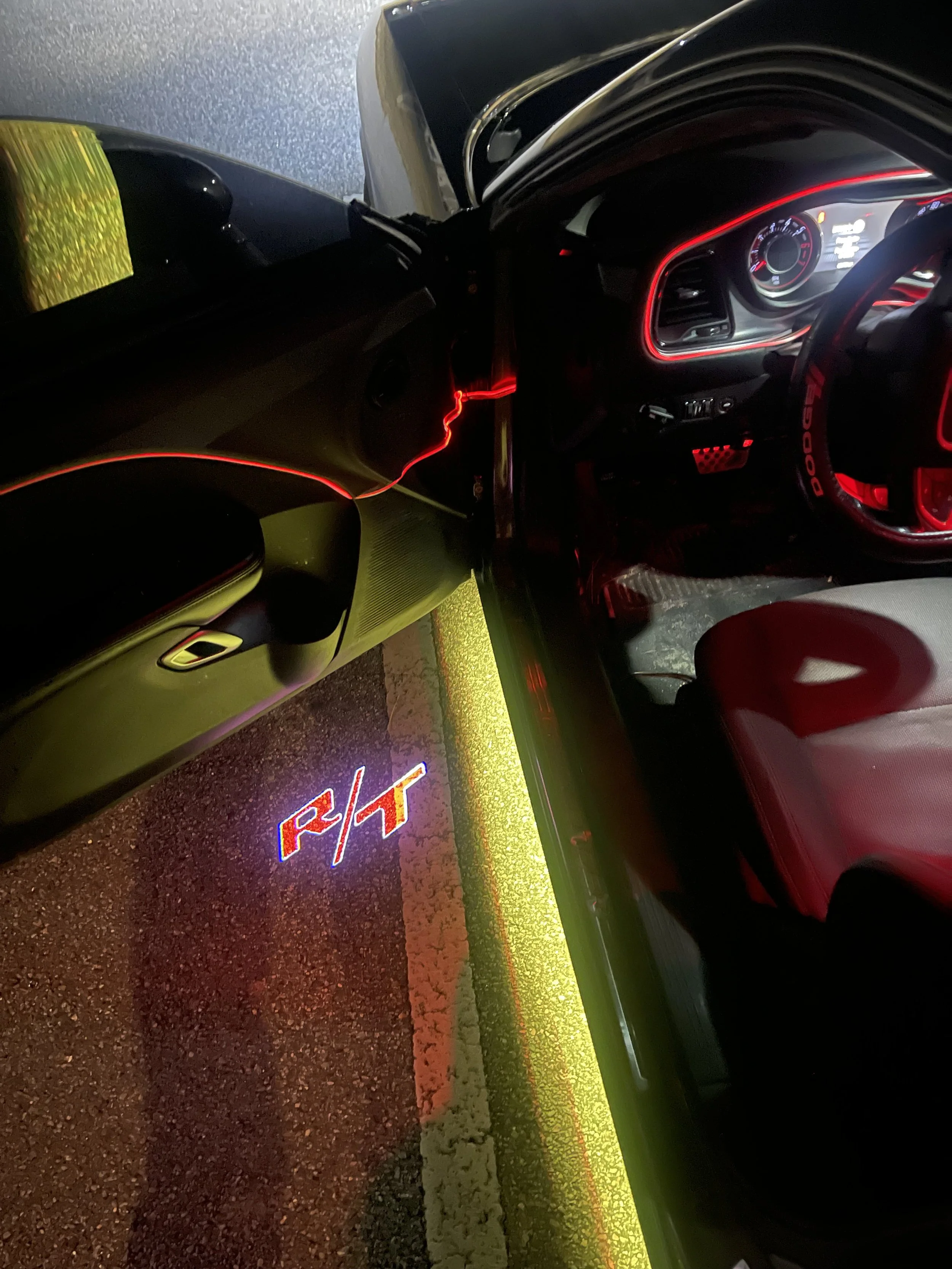 Nighttime view of a car with red neon lights outlining the dashboard and interior, parked outside near a sign with illuminated red and yellow text on the ground.