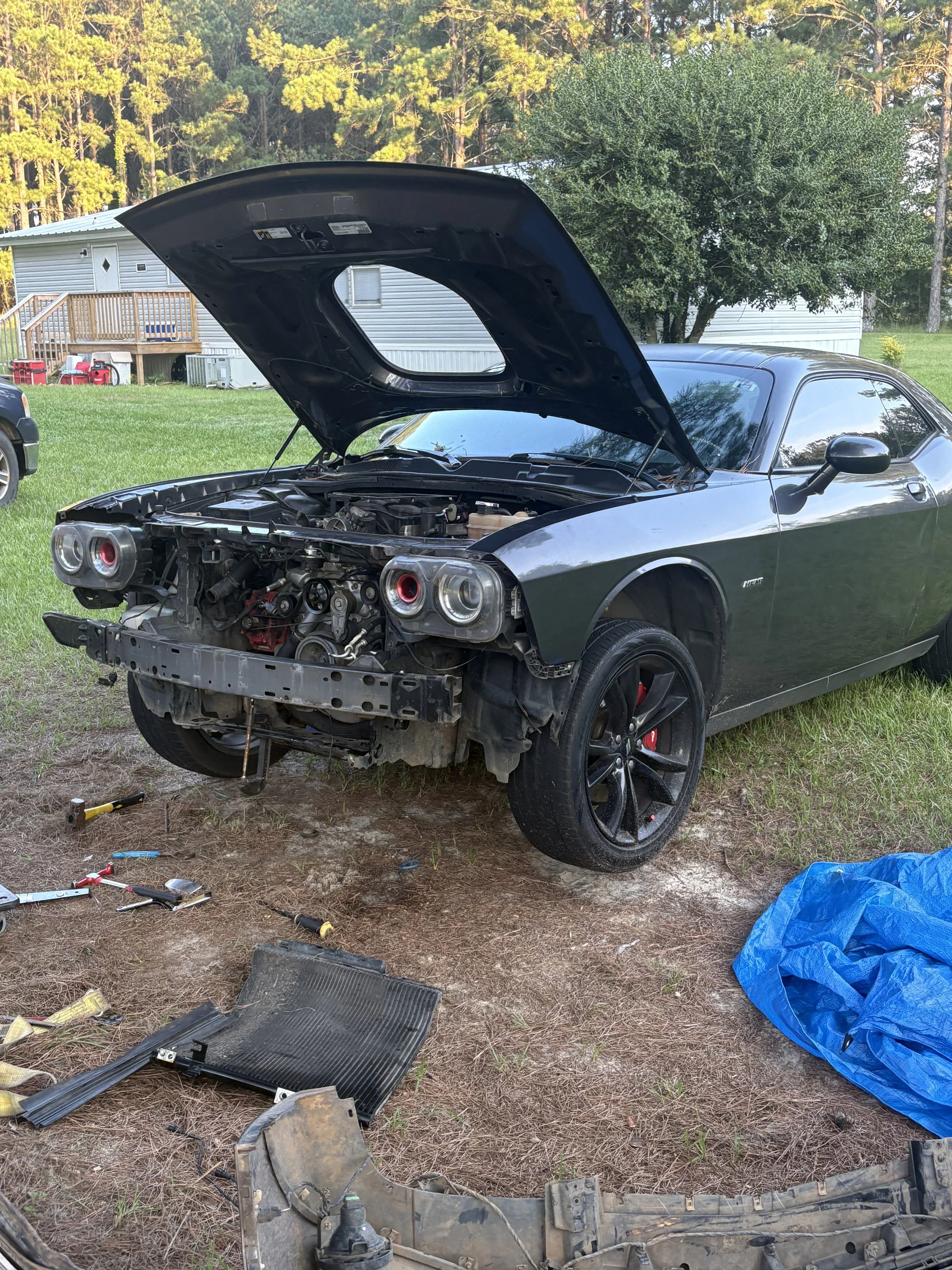 A black sports car with its front bumper and parts removed, parked on grass. The hood is open, revealing the engine. Tools and car parts are scattered on the ground nearby, with a blue tarp on the right side. In the background, there is a house with 