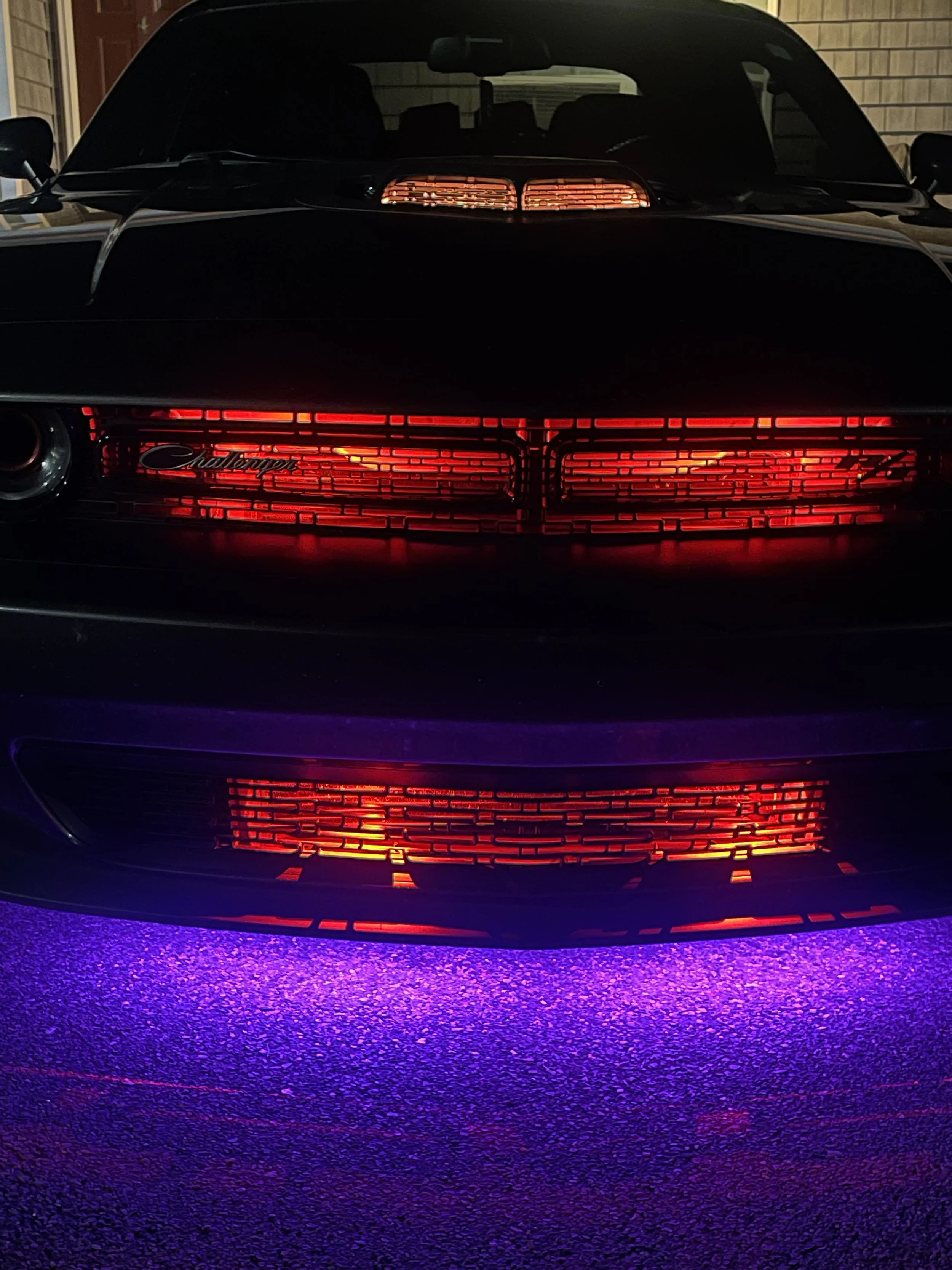 The front of a black car with illuminated red grille and taillights, parked on a purple-lit asphalt surface at night.