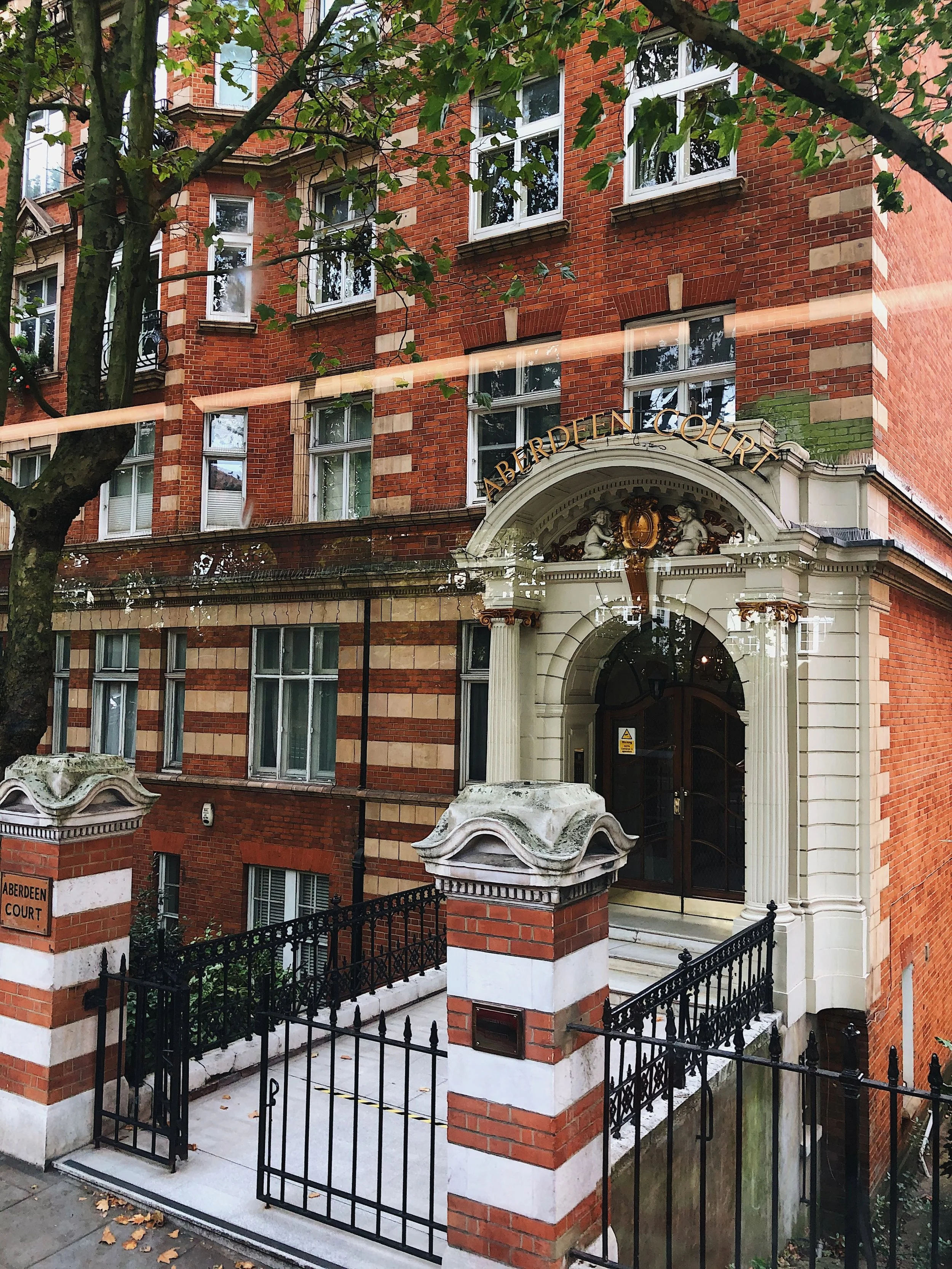 View of a red brick building with multiple windows, an ornate entrance arch, and a black metal fence around the entrance. A tree with green leaves partially obscures the view.