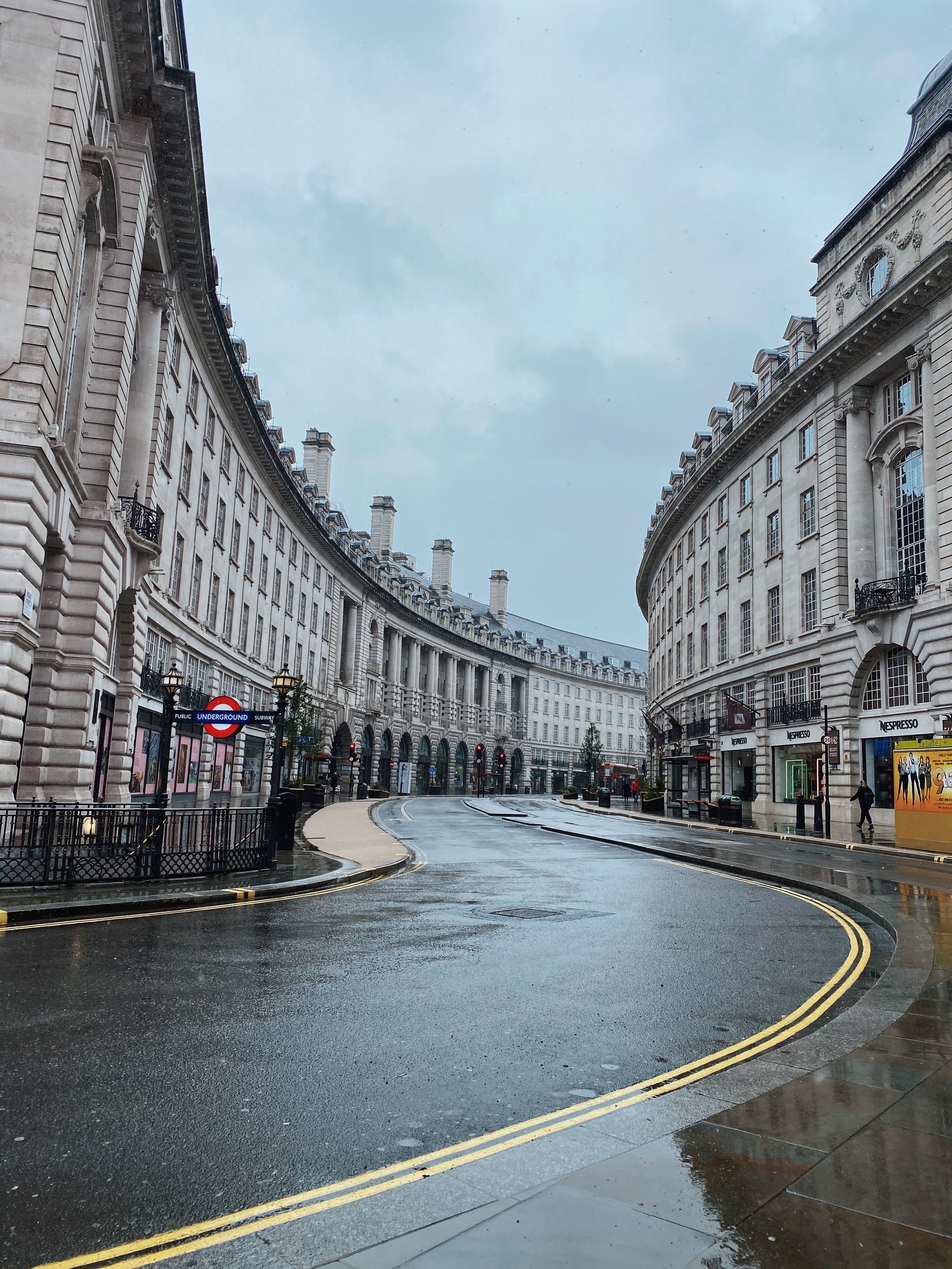 Rain-soaked street in a European city with classic white stone buildings, a bus stop with an Nespresso advertisement, and an underground subway sign. The street is curved with wet pavement and yellow double lines, and some pedestrians are visible.