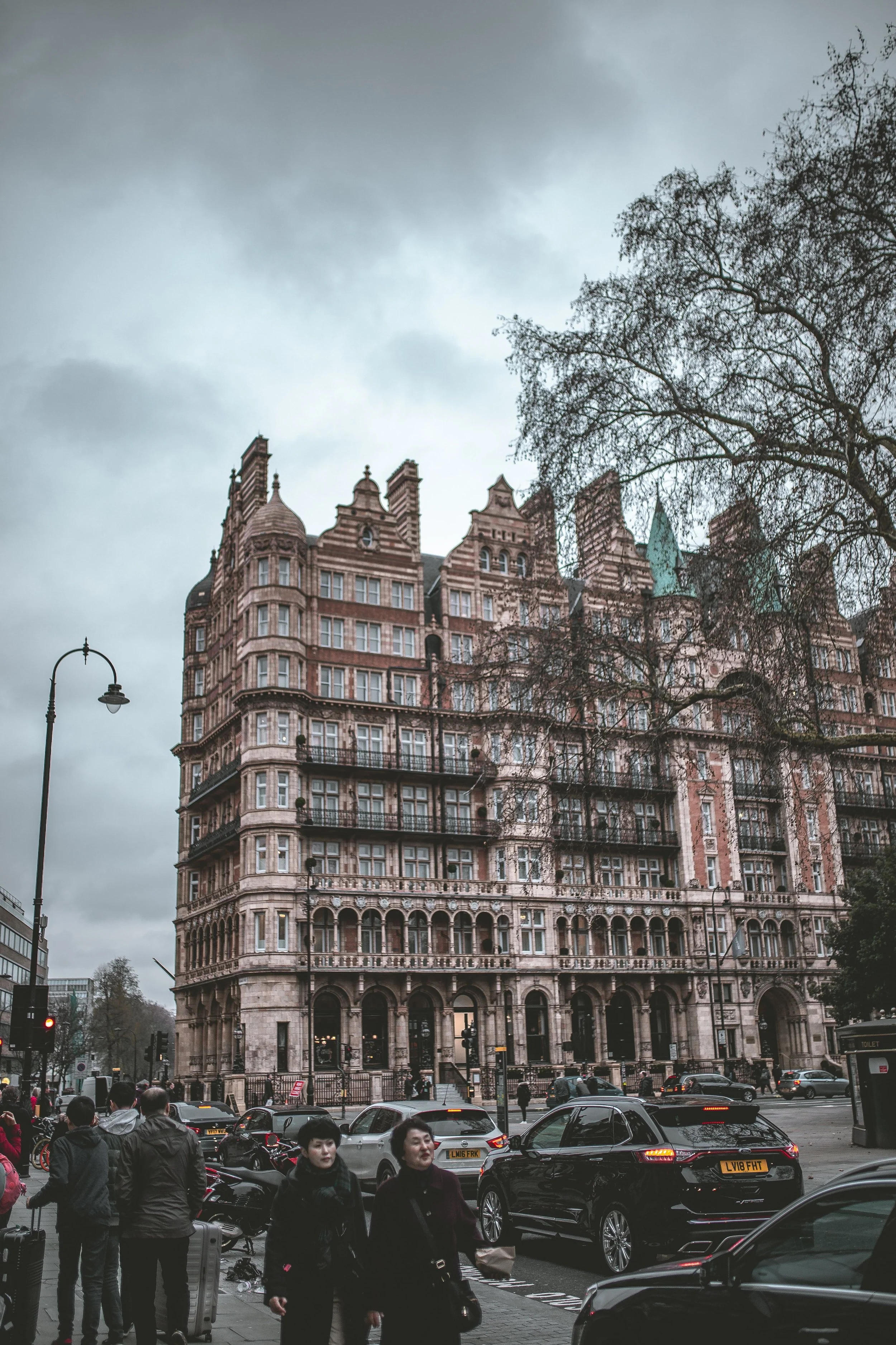 A busy city street with people walking and various cars parked and driving. In the background, there is a tall, ornate, pinkish historic building with intricate architecture, and a cloudy sky overhead.