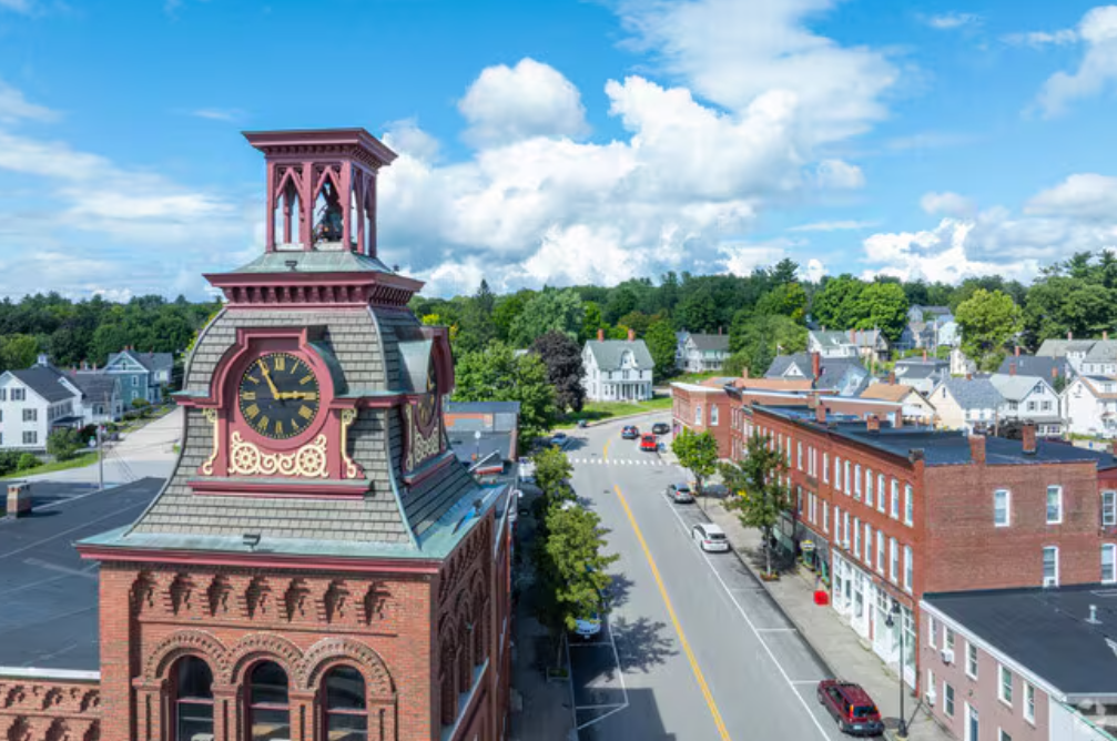 Aerial view of a small town with a prominent brick clock tower, streets lined with trees and cars, and residential houses in the background under a partly cloudy sky.