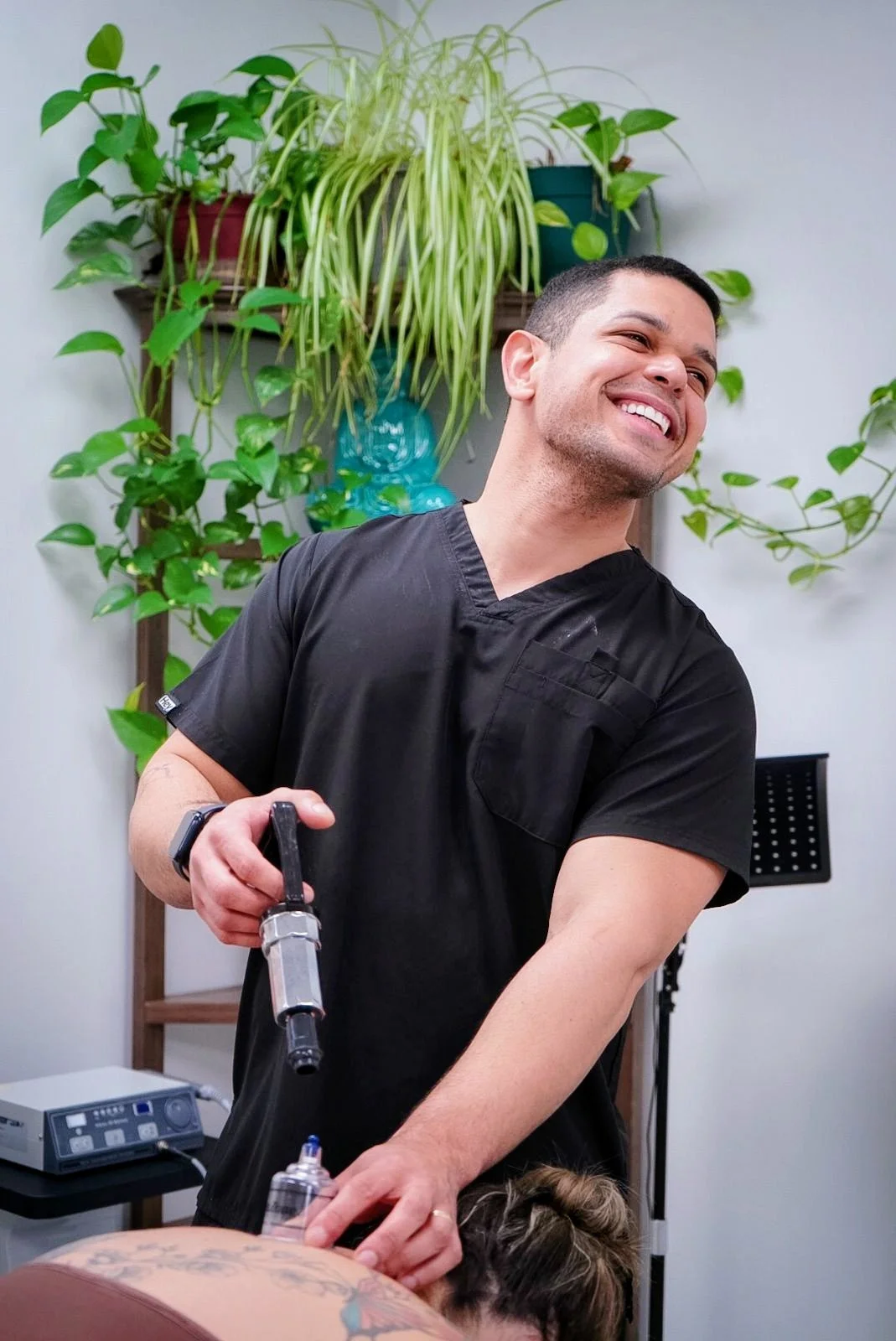 A man smiling while preparing to inject a tattoo into someone's arm, with green plants on the wall behind him.