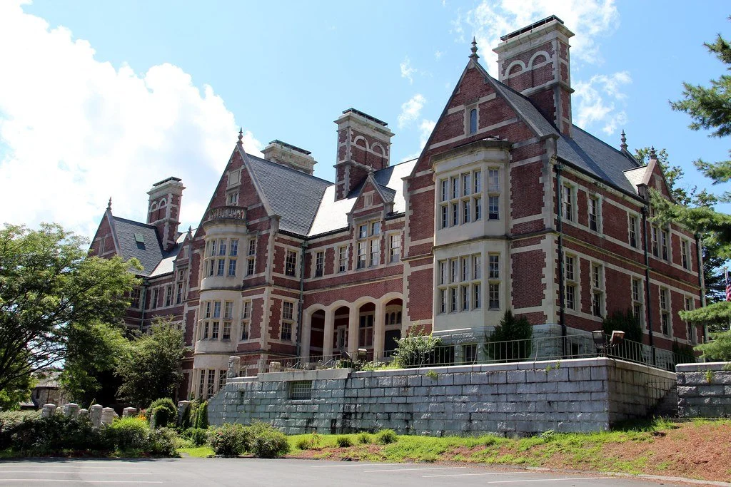 Large historic brick mansion with multiple turrets and elaborate architecture, surrounded by greenery and a stone retaining wall.