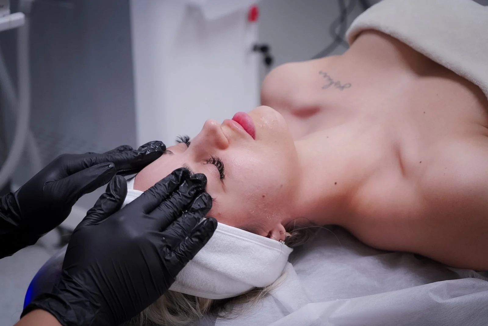 A person receiving a facial treatment at a spa or clinic, lying with eyes closed, with a skincare professional wearing black gloves applying treatment to their face.