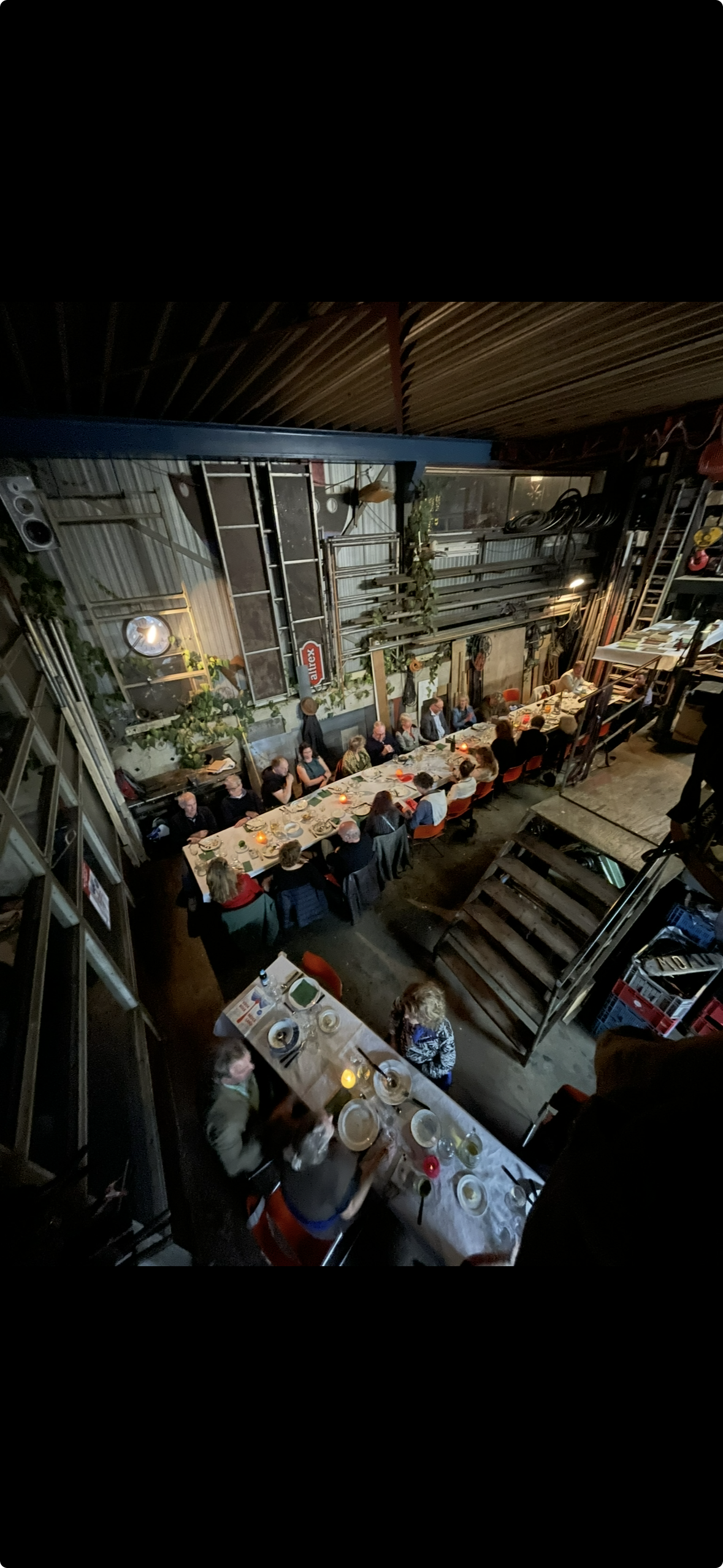 Gezellige groep mensen zit aan een lange eet- en drinktafel in een industrieel restaurant of café, met kaarslicht en planten, binnen een ruime, open structuur.