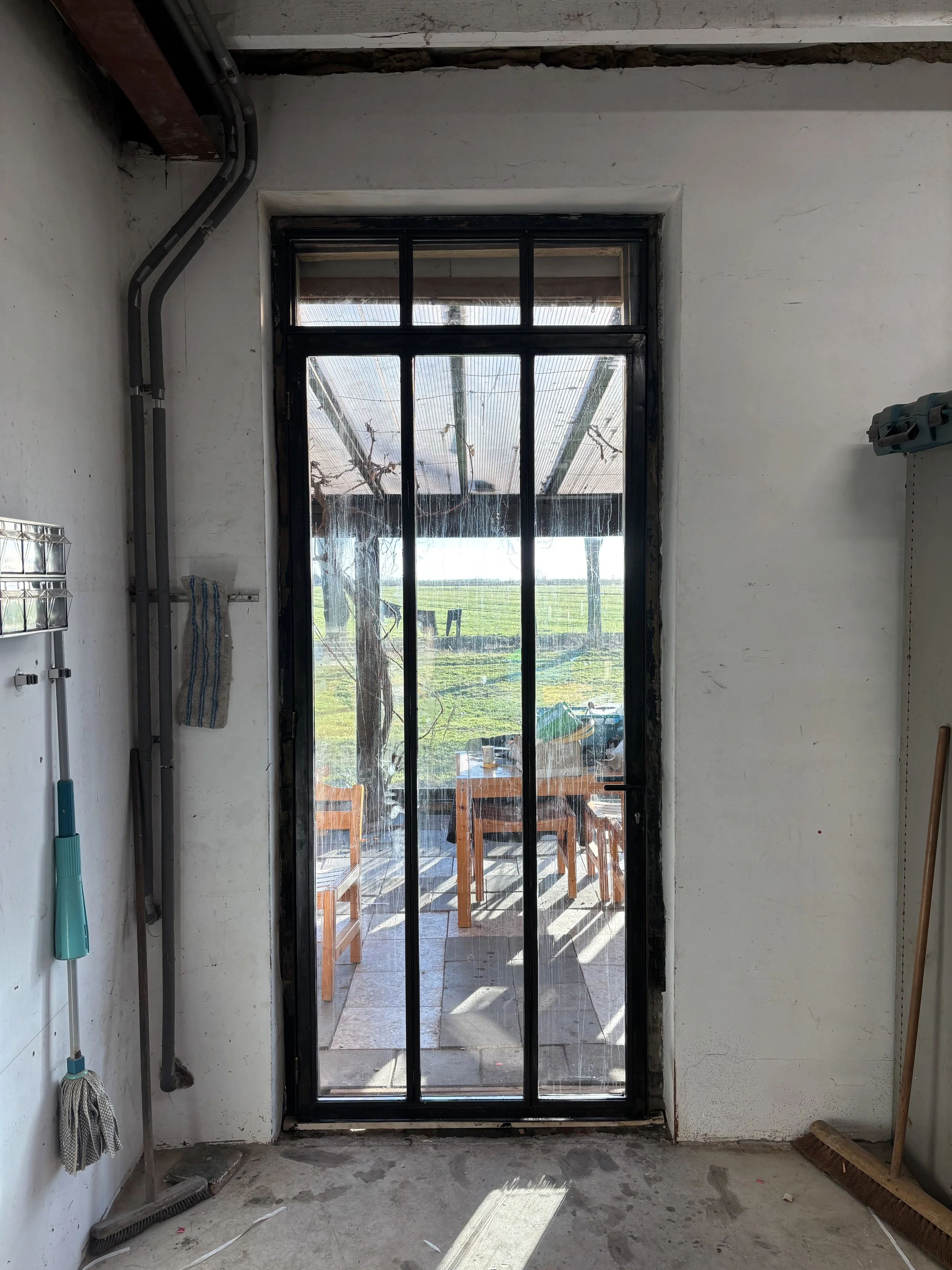 Binnenkant van een kamer met een zwarte metalen deur die uitkijkt op een terras met tuinmeubels en uitzicht op een groen veld. De kamer is niet afgewerkt en bevat schoonmaakgereedschap en een uitgestreken muur.