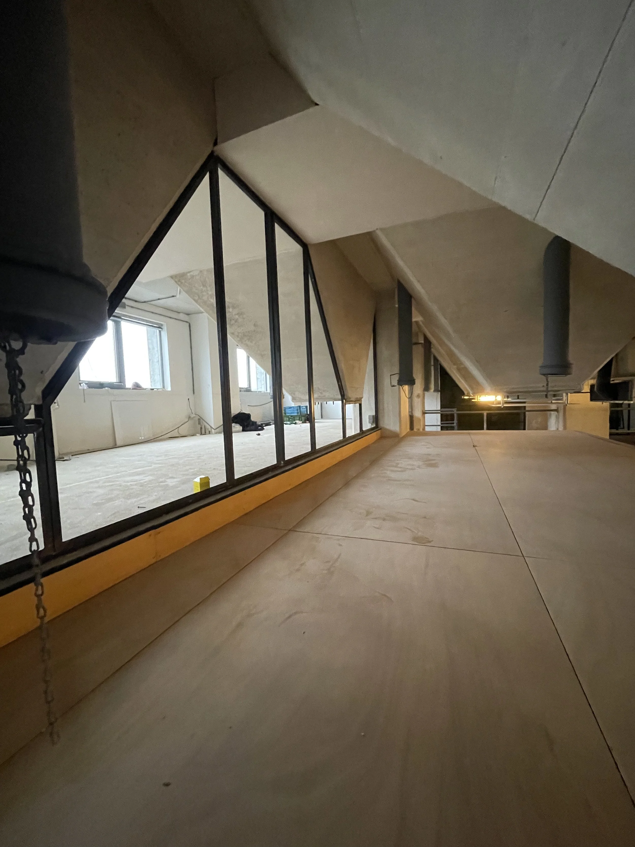 Interieur van een zolder met onafgewerkte wanden, stukke hout en bouwmaterialen, ramen en een enkele lamp, mogelijk in aanbouw of renovatie.