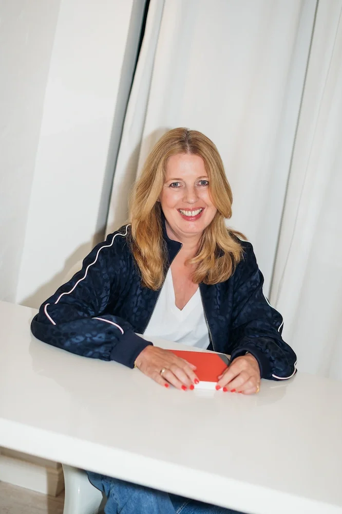 Birgit Drixelius sitzt lächelnd an einem weißen Tisch und hält ein rotes Buch.