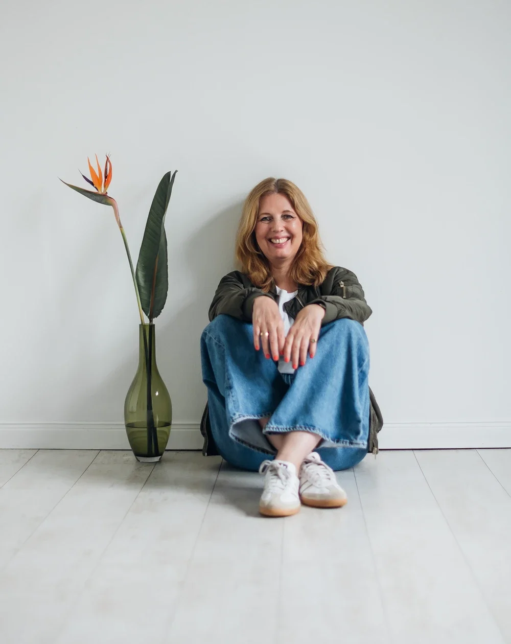 Birgit Drixelius lächelt in die Kamera. Sie sitzt lässig auf dem Fußboden. Neben ihr eine Vase mit einer exotischen Blume.