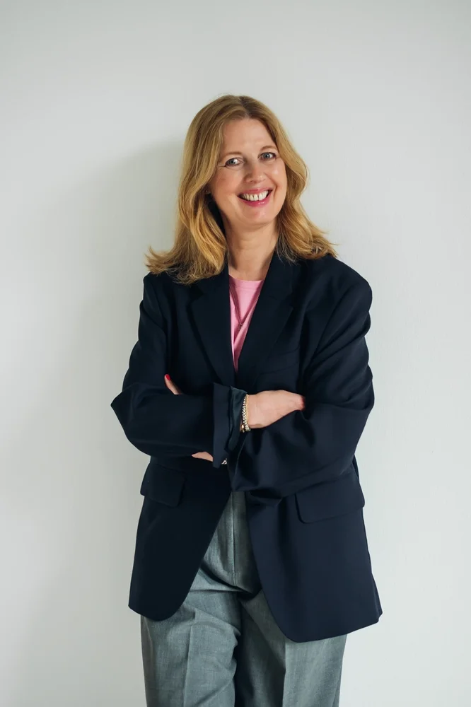 Profilbild von Birgit Drixelius - Beraterin für FührungskulturSie trägt eine graue Hose, rosa Hemd und dunkler Jacke, vor weißer Wand.