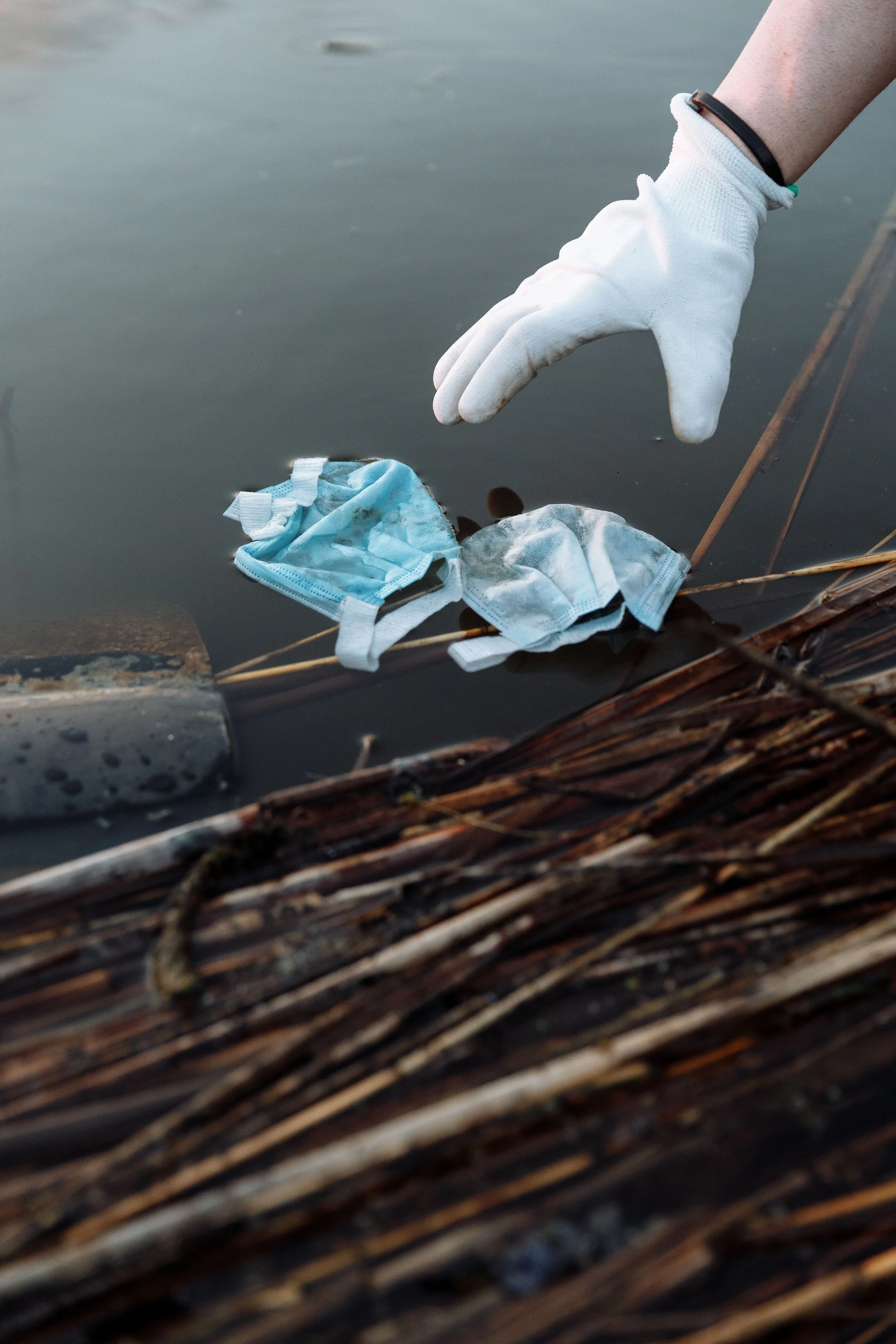 A pair of disposable face masks on a surface near the edge of a body of water, with a gloved hand reaching towards them.