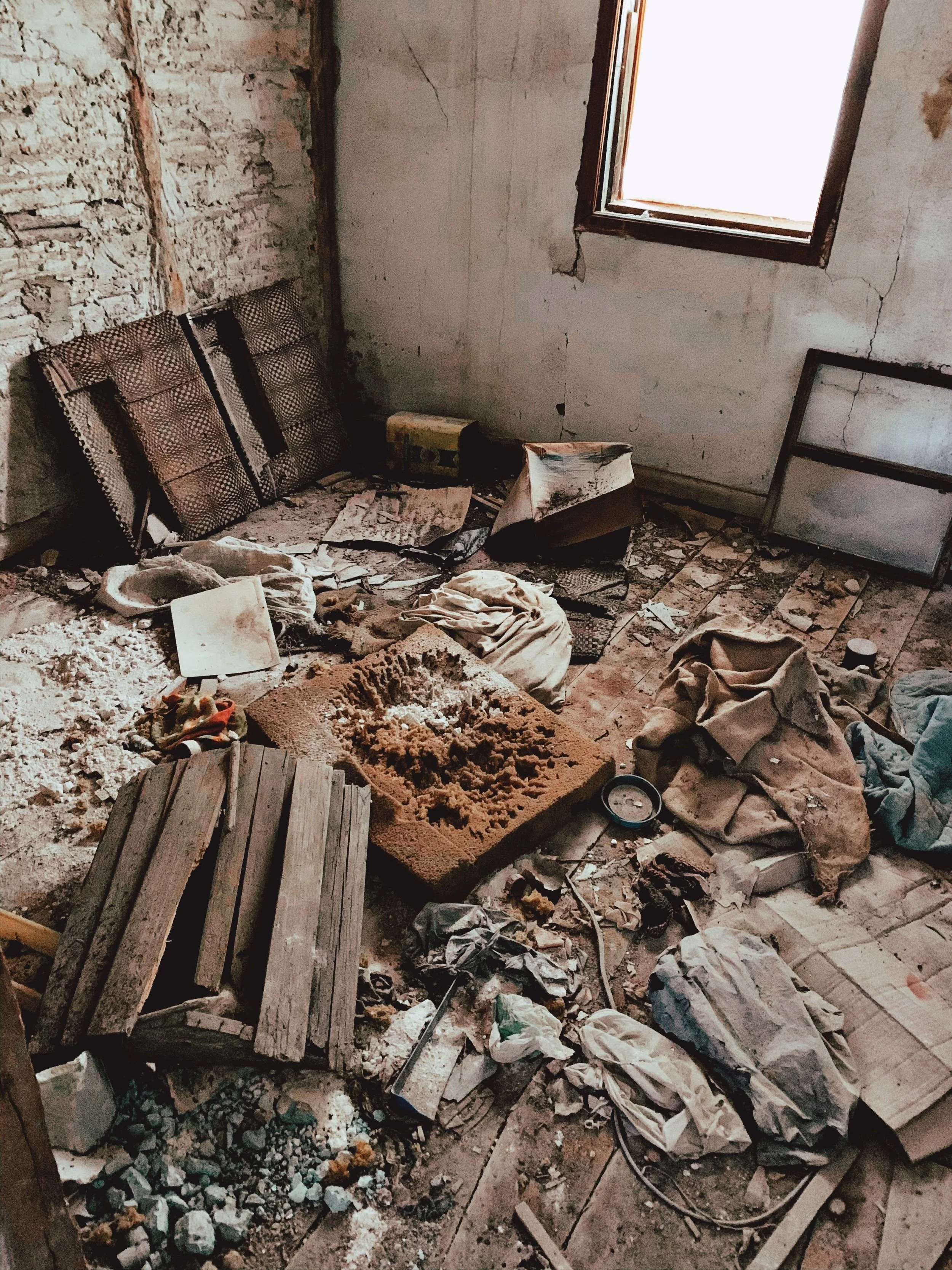 An interior room in disrepair with exposed brick and plaster walls, a single small window, scattered debris, rocks, and old furniture, including a broken chair and a wooden pallet.