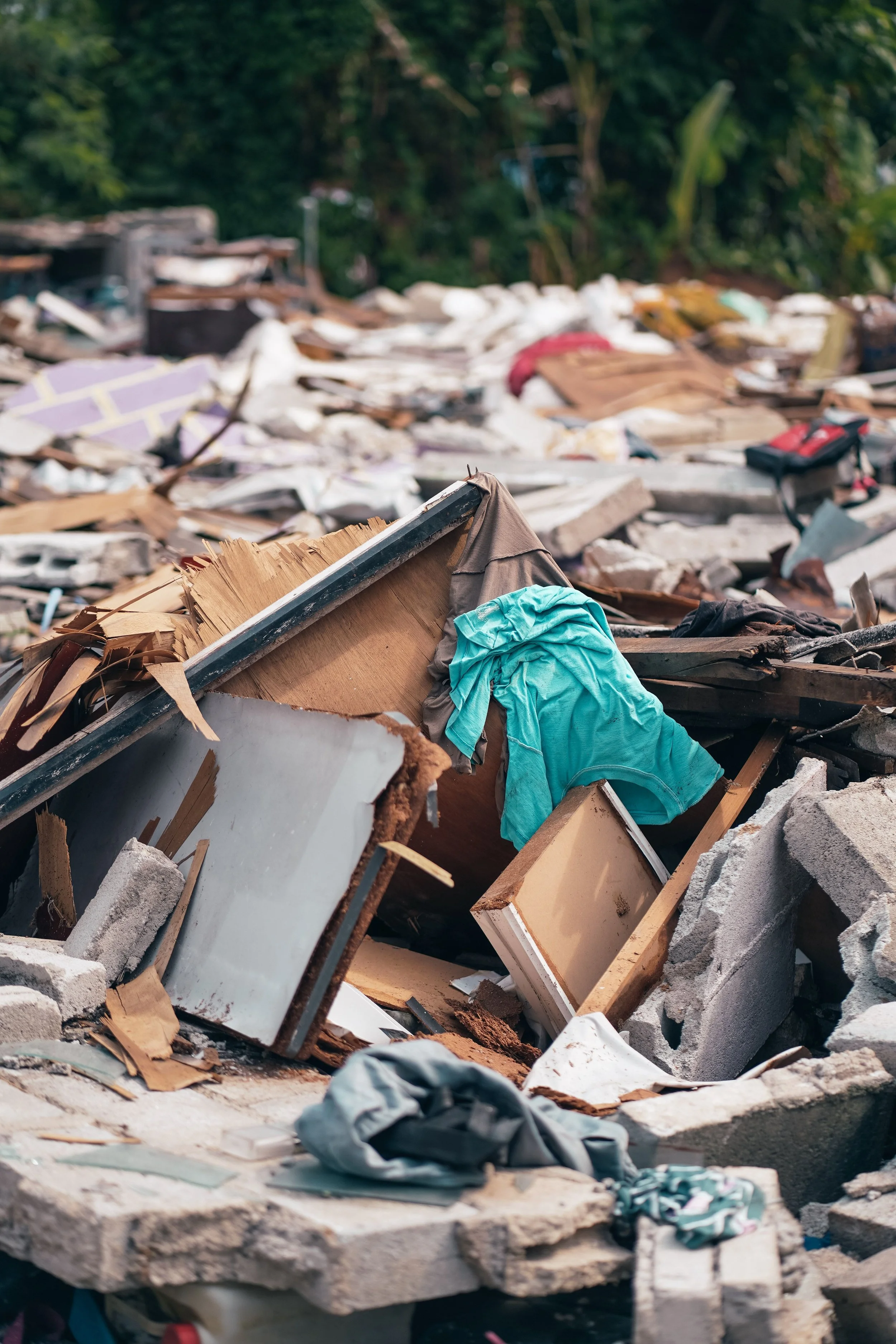 A large pile of debris including broken furniture, household items, and rubble, with clothing and fabric scattered on top, set outdoors with green trees in the background.