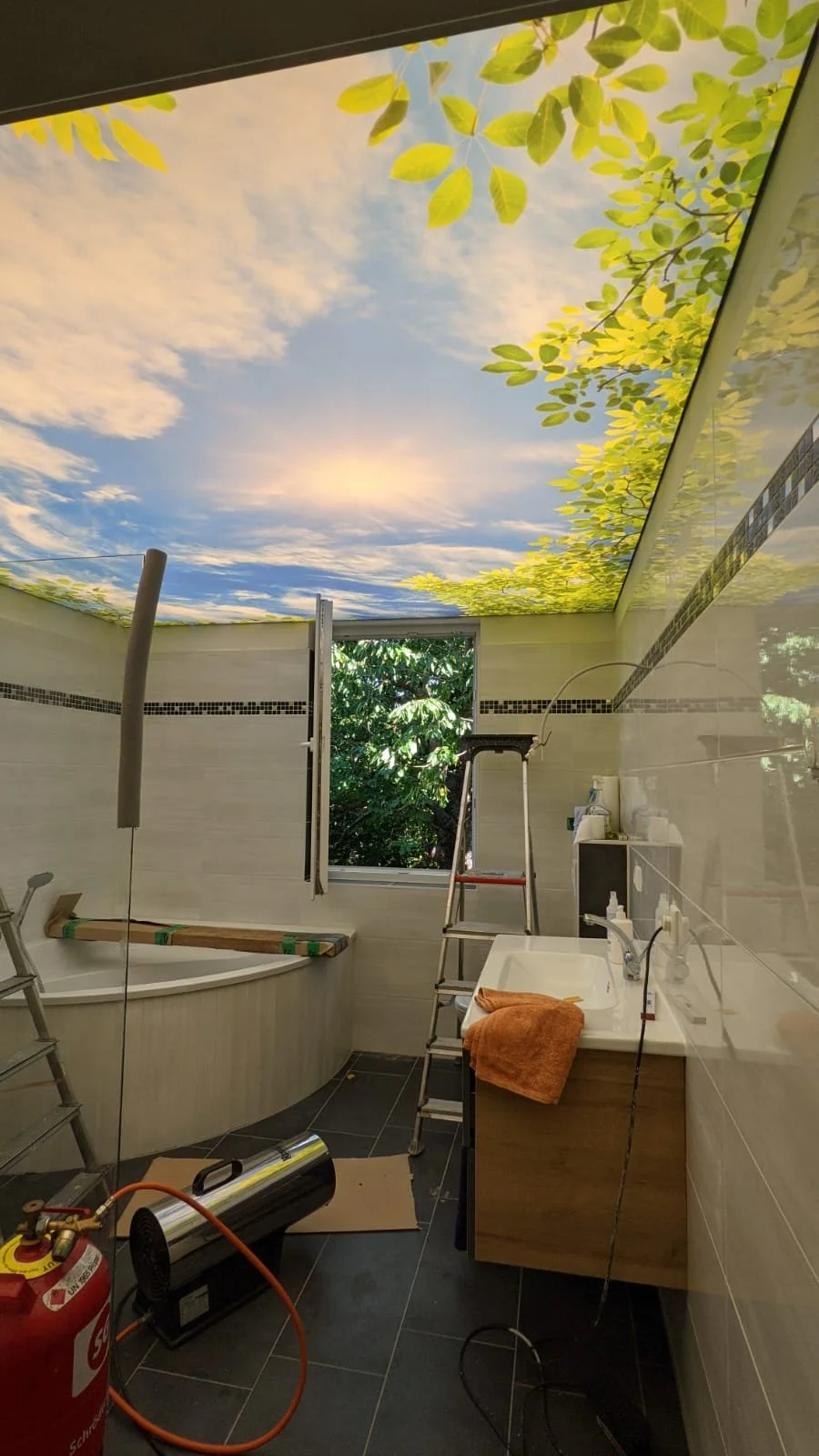 Innenansicht eines Badezimmers mit geöffnetem Fenster, das einen grünen Baum und bewölkten Himmel zeigt, Decke mit Himmel- und Baum-Motiv, Badewanne, Waschbecken, Leiter und Reinigungsgeräte.