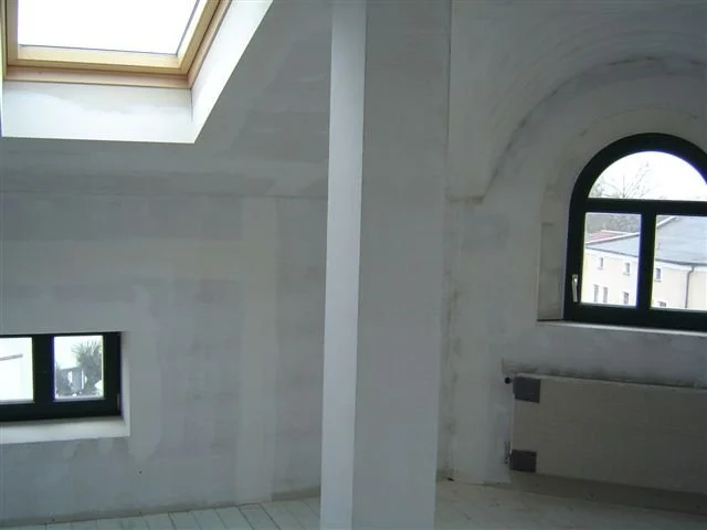 Innenaufnahme eines Raums mit Dachfenster und einem Bogenfenster, helle Wände und Boden, keine Möbel sichtbar.