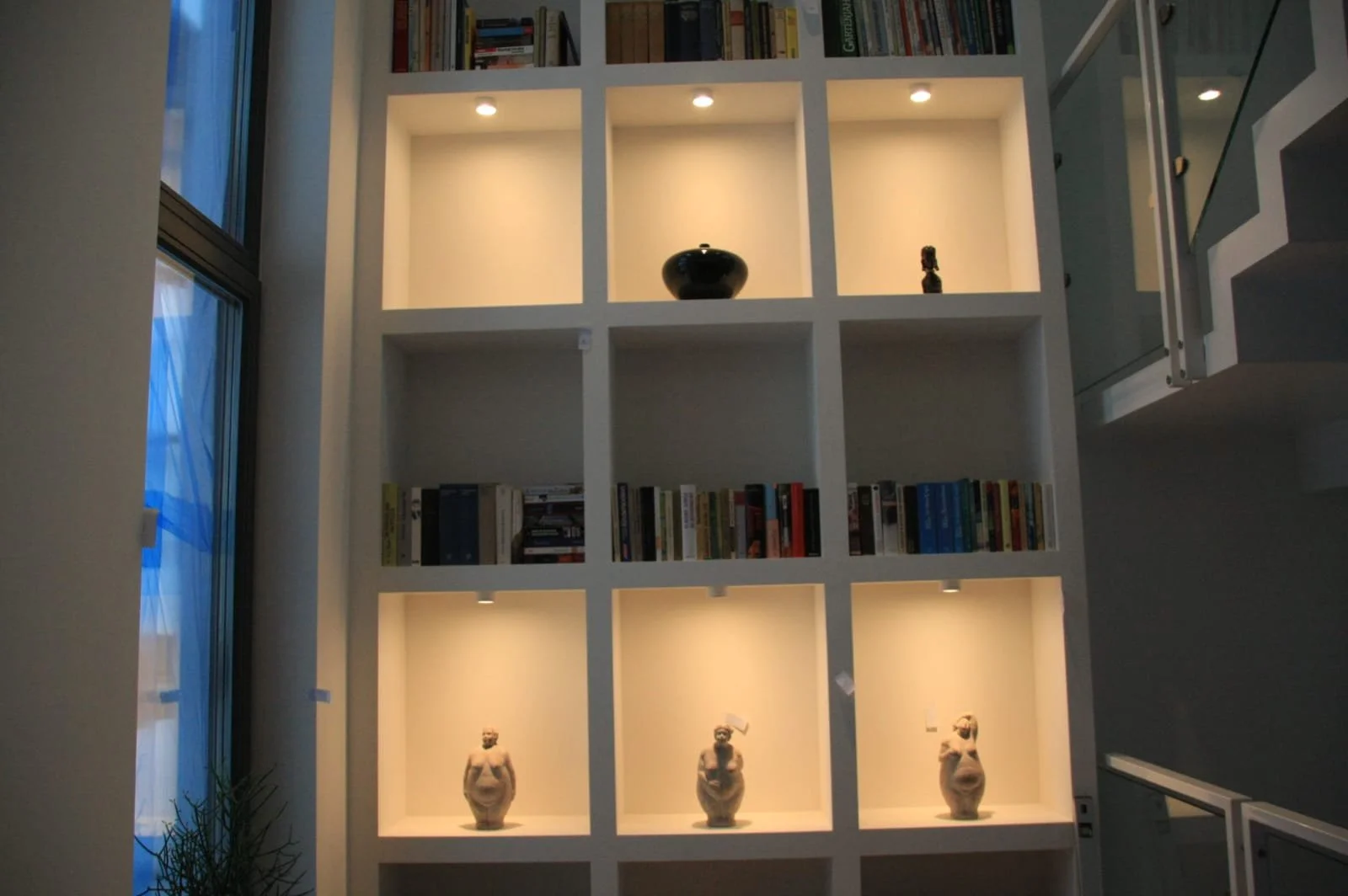 Eine weiße, in quadratische Fächer unterteilte Bücherregal mit Beleuchtung, das auf einem erhöhten Boden steht. Es enthält Bücher und dekorative Figuren, darunter drei Skulpturen am unteren Rand.