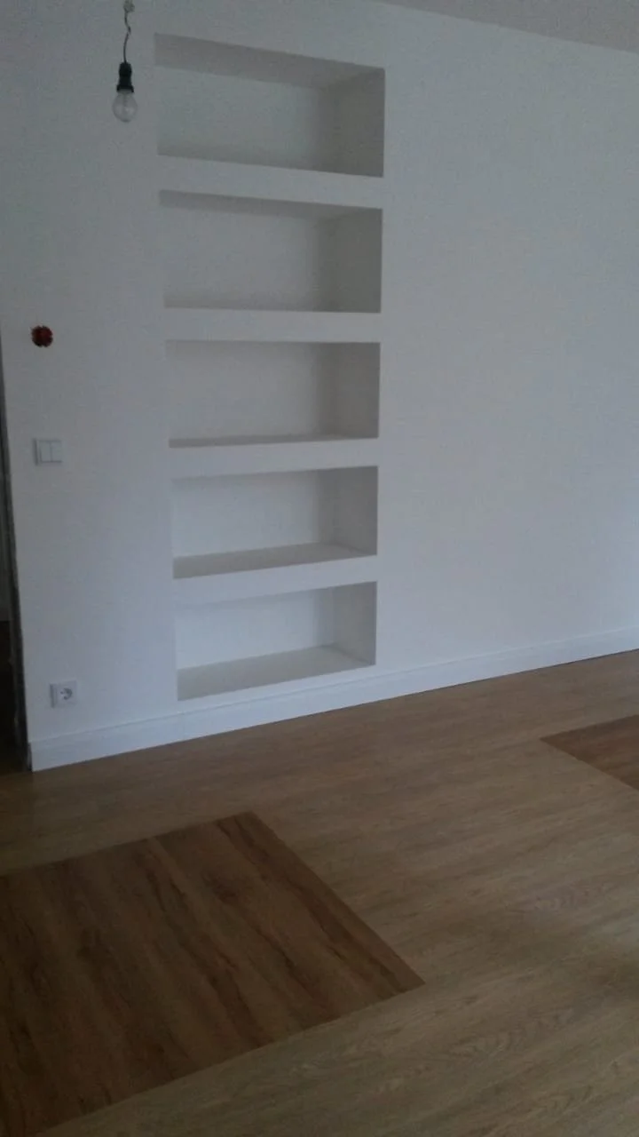 Leere weiße Wand mit eingebauten Nischen in einem Raum, Holzboden, im oberen Teil eine offene Glühbirne ohne Lampenschirm, und ein rotes Element, möglicherweise ein Rauchmelder, auf der linken Wandseite.