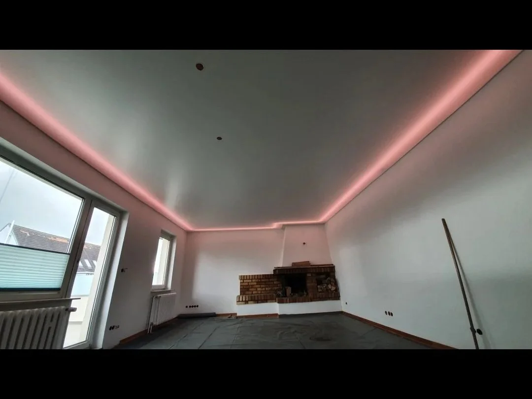 Leeres Wohnzimmer mit recent renovierten weißen Wänden, einem Kamin aus Ziegeln, großen Fenstern und einer Deckenbeleuchtung in Pink.