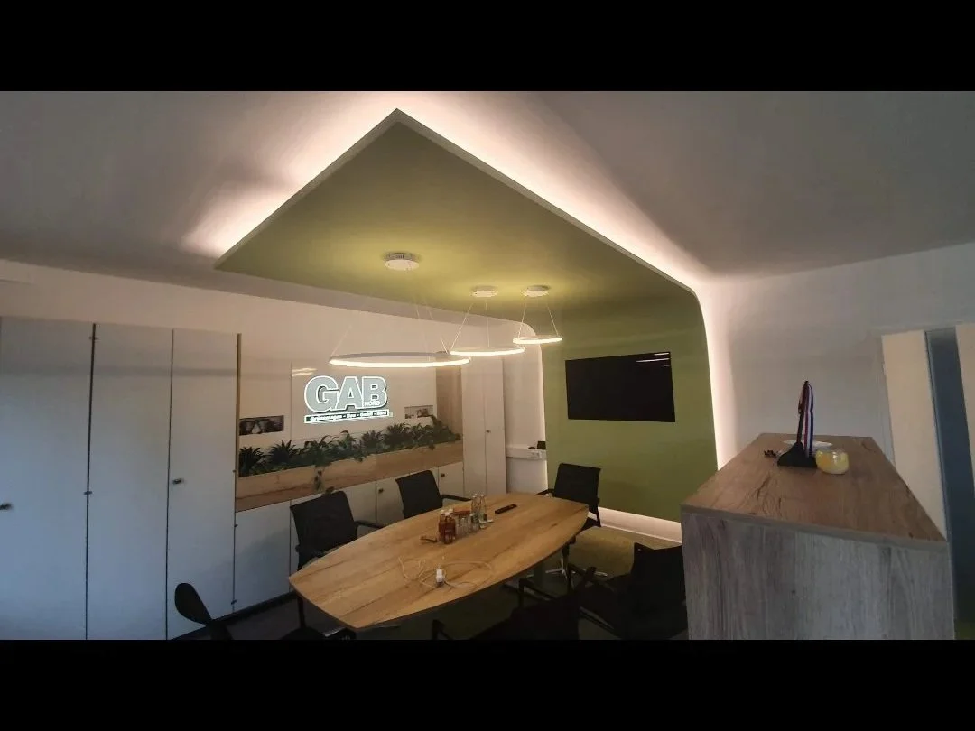 Ein moderner Conference-Raum mit einem großen Holzkonferenztisch, schwarzen Stühlen, einem grünen Akzentwand mit einem Fernseher und einer Deckenleuchte, sowie einem Whiteboard mit dem Logo 'GAB' auf einer Wand.