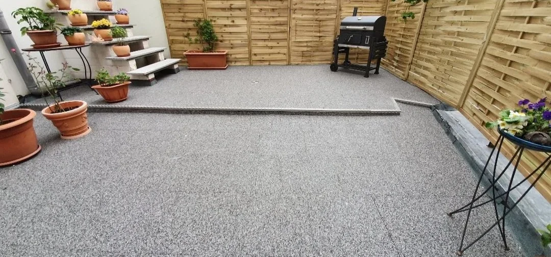 Gepflasterter Gartenbereich mit Pflanzkübeln, einer kleinen Treppe, einem Grill und einem Blumentopf mit Blumen.
