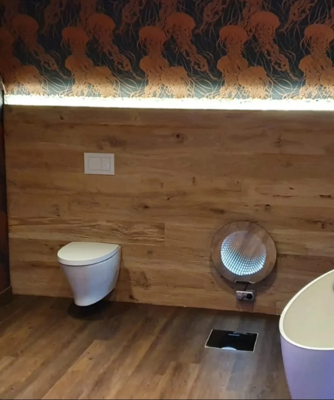 Ein Badezimmer mit Holzverkleidung, einer modernen wandhängenden Toilette und einem runden Waschtisch. Es gibt auch eine runde Lüftungseinrichtung an der Wand.