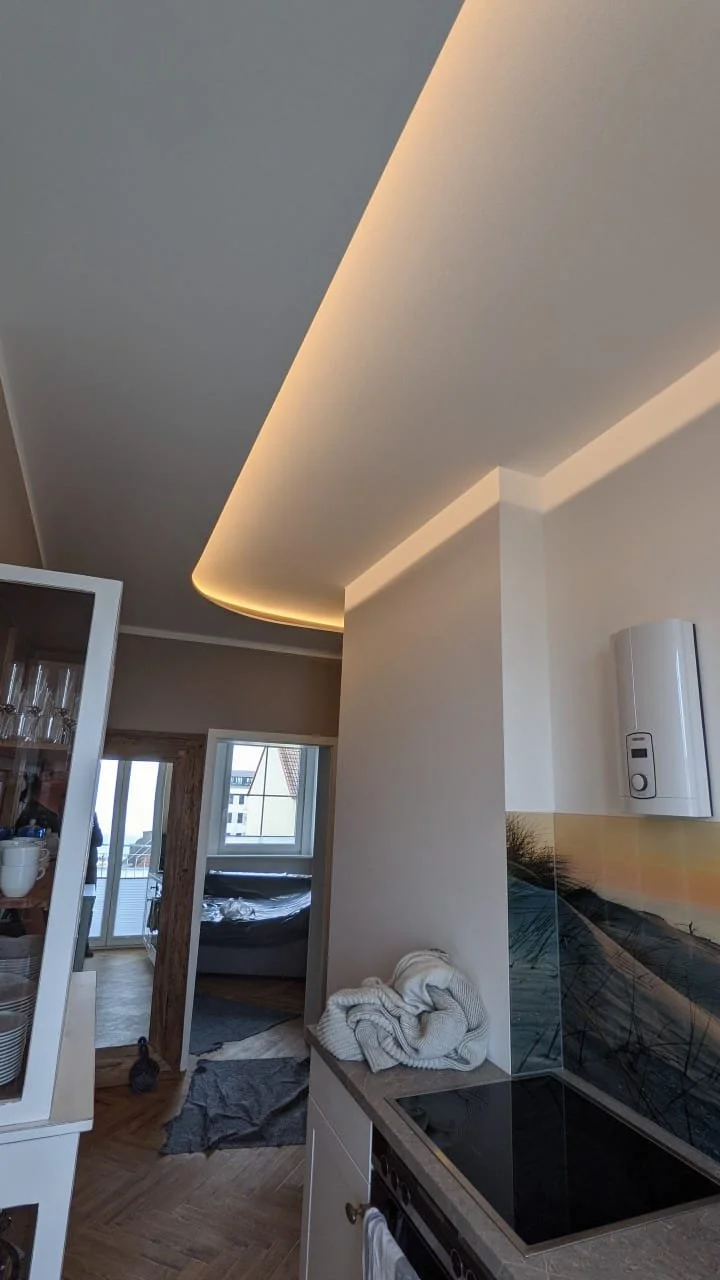 Innenansicht einer modernen Wohnung mit sichtbarem Küchenbereich, offener Tür zum Schlafzimmer, Garderobe und Balkon, gedämpftes Licht an der Decke.