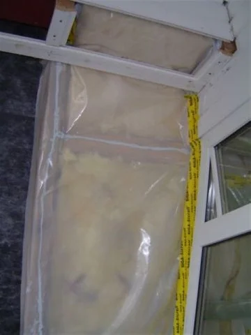 Was scheint eine verschlossene Plastiktüte mit einem weißen, verschmutzten Gegenstand darin vor einem Fenster, daneben ein gelbes Maßband.