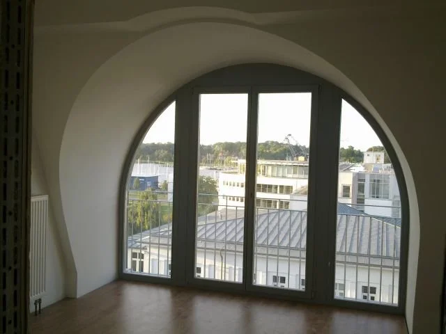 Blick durch ein großes, gebogenes Fenster in eine Stadt mit modernen Gebäuden und Baumflächen im Hintergrund.