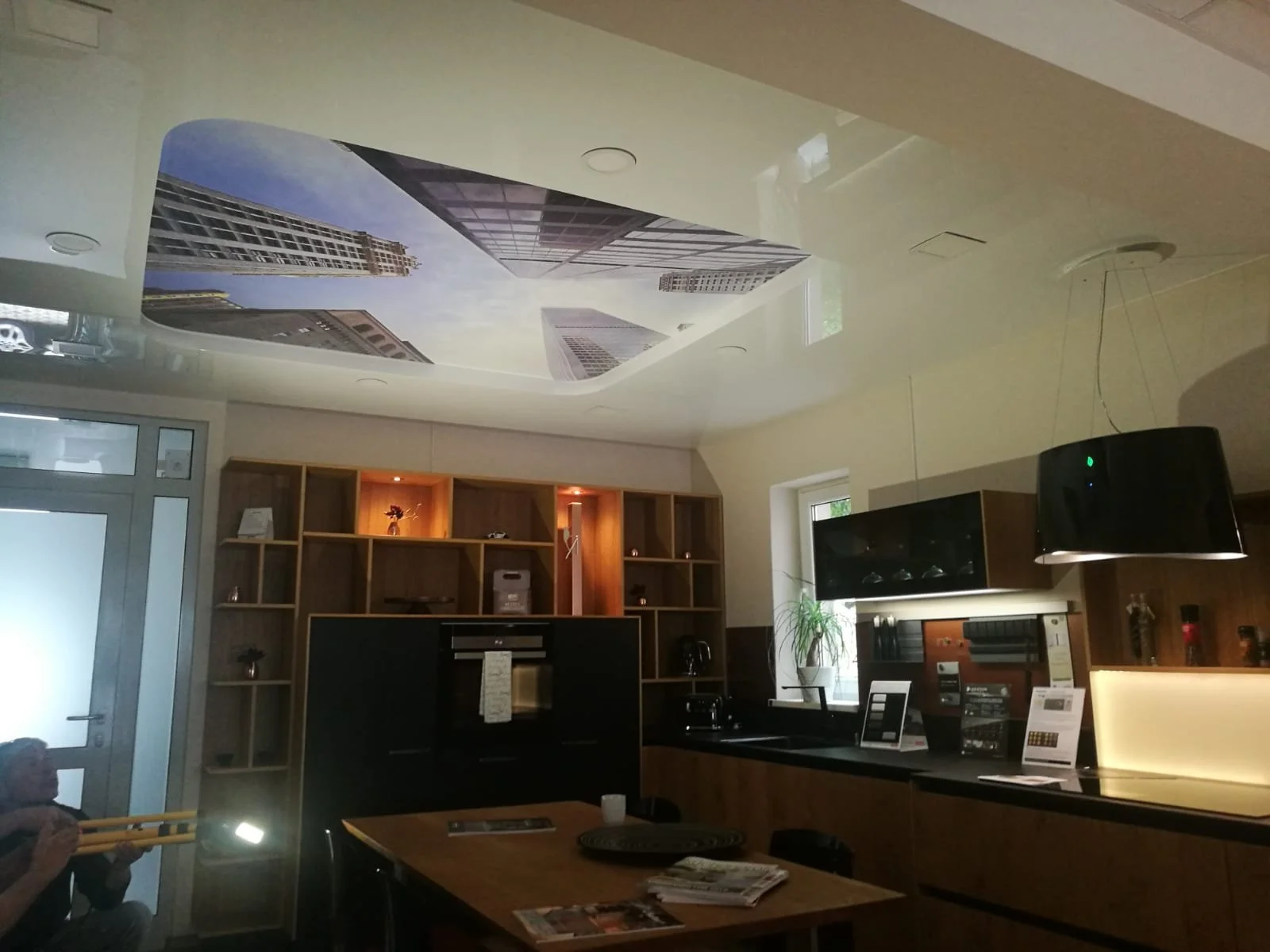 Decke mit großem Bild von Wolkenkratzern in urbaner Umgebung, Wohnzimmer mit Holzregalen, Fernseher, Tisch mit Zeitschriften und Personen, die entspannt sitzen.