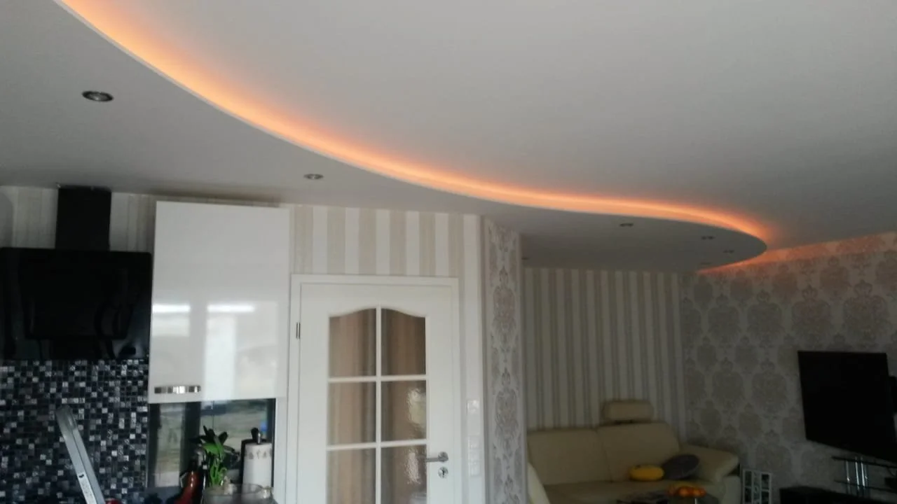 Ein modernes Wohnzimmer mit einer Deckenleuchte, einer weißen Tür, einem Fernseher, einem Sofa und einer Wandverkleidung.