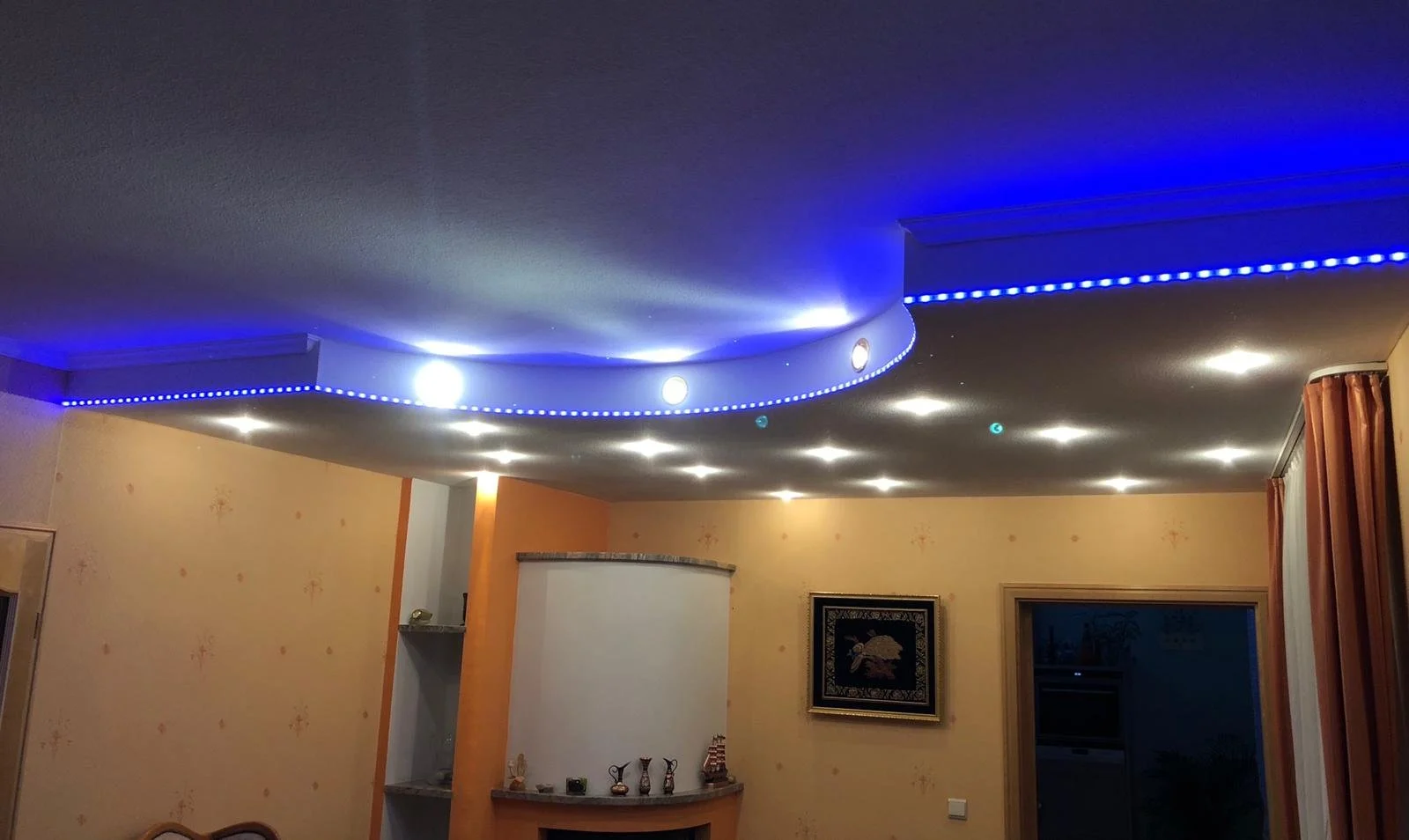 Innenraum mit Deckenbeleuchtung, blauer LED-Leiste und Dekorationsgegenständen auf einem Regal über dem Kamin.