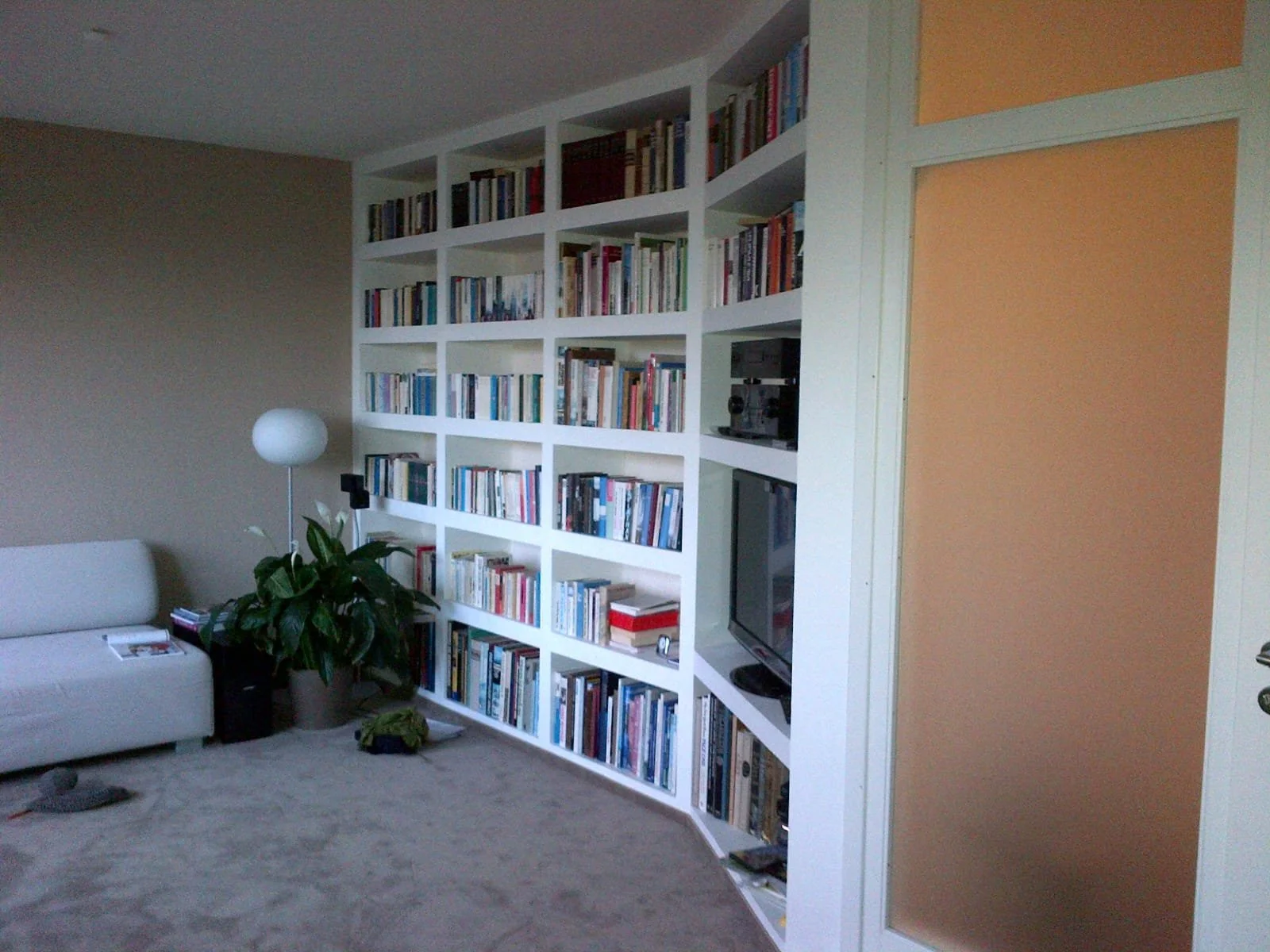 Wohnzimmer mit weißem Sofa, einer großen Bücherwand, einem Pflanzen, einem Stehlampe, Fernseher und einem beige-hellblauen Wand. Es gibt auch eine Zimmerpflanze und Zeitschriften auf dem Sofa.
