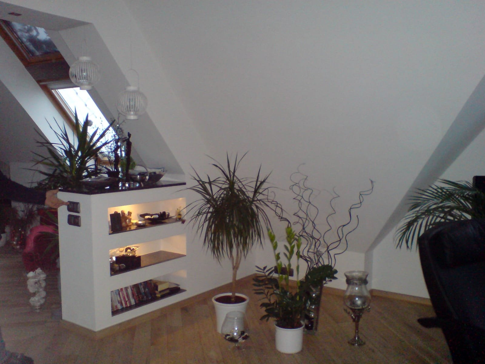 Innenraum mit mehreren Topfpflanzen, einem weißen Bücherregal, Kerzen und dekorativen Gegenständen, im Hintergrund eine Dachschräge mit Fenstern.