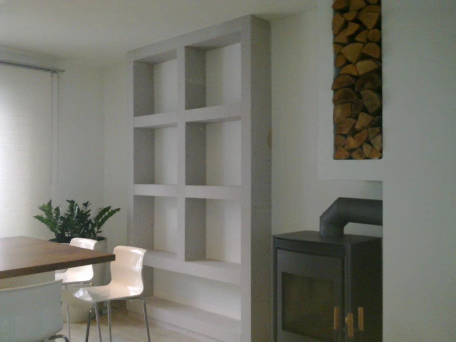 Ein weißer Bücherregal ohne Bücher an einer weißen Wand in einem Wohnzimmer, daneben ein Holzofen, darüber Brennholzstapel, im Vordergrund Teil eines Esstisches mit weißen Stühlen.