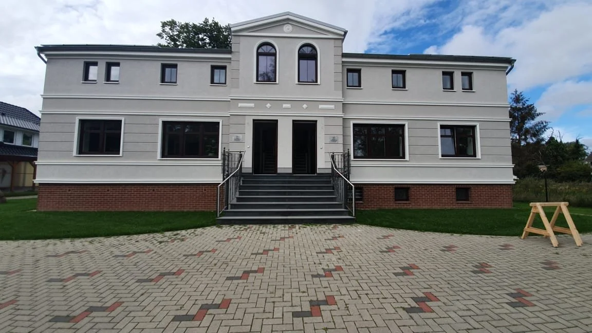 Geräumiges, zweistöckiges, weißes Haus mit einem zentralen Eingang, Treppe und mehreren Fenstern, umgeben von einem gepflasterten Hof und Grünfläche.