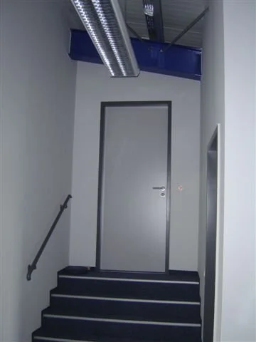 Eine geschlossene Tür am Ende eines Treppenhauses mit schwarzen Stufen und weißen Wänden.