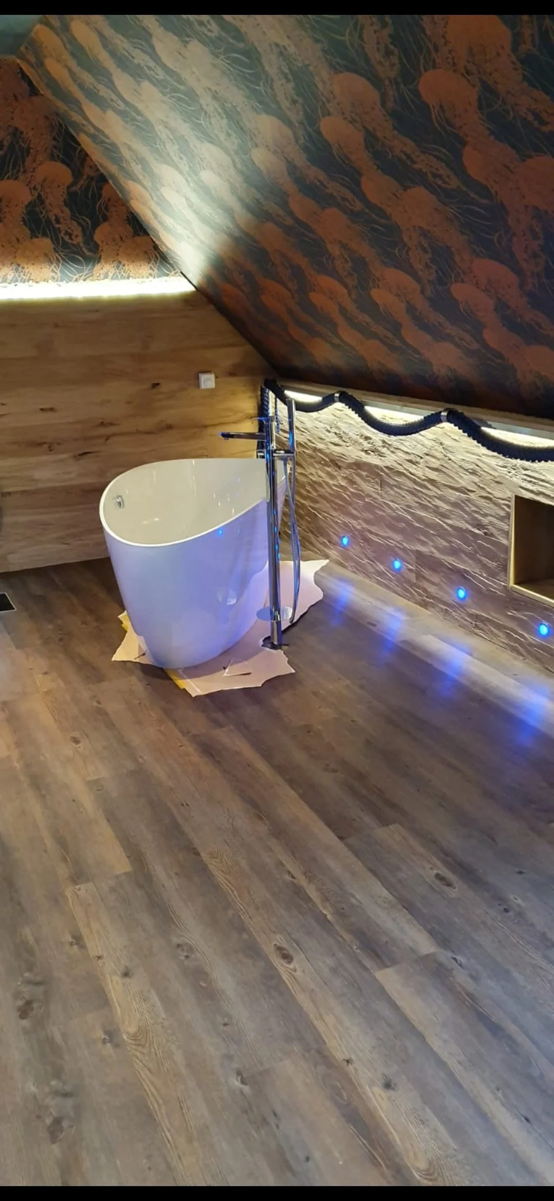 Eine moderne Badewanne mit freistehender Badarmatur in einem Raum mit Holzboden und Wandverkleidung aus Holz und Stein, dekorativ beleuchtet mit blauen LEDs und einer Wandverzierung mit Muschelmuster unter einer schrägen Decke.