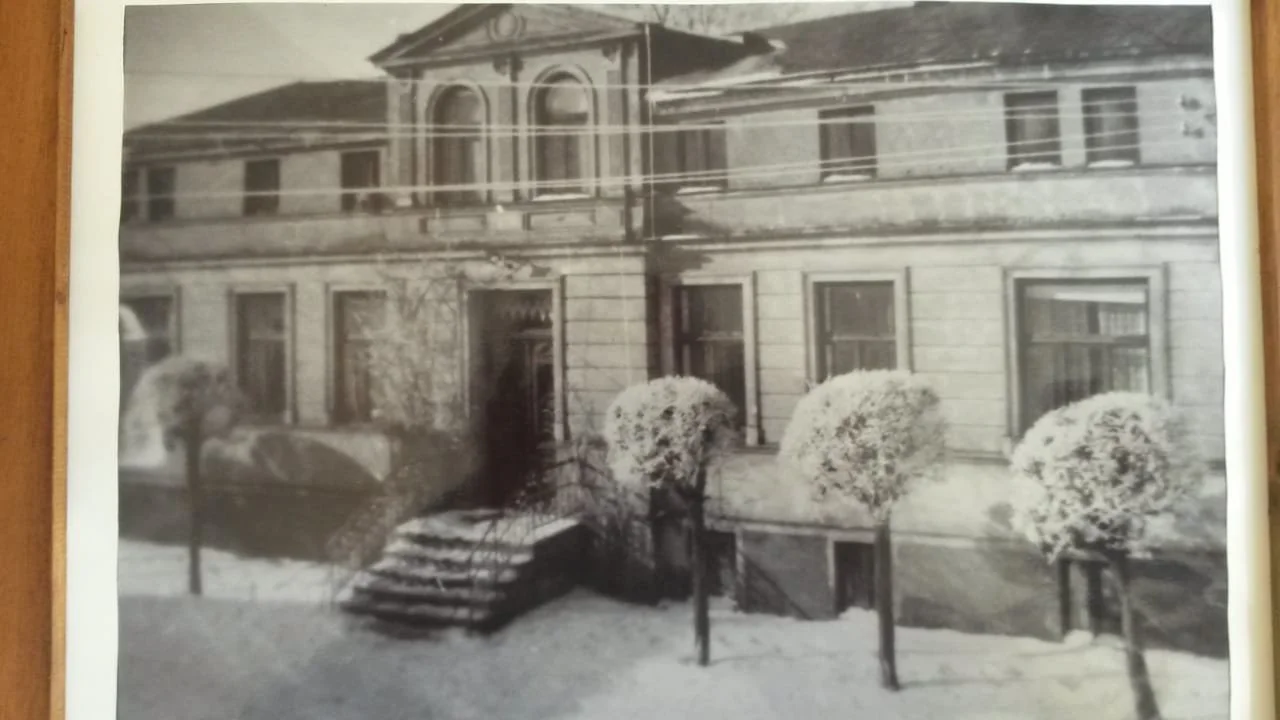 Schwarz-Weiß-Foto eines historischen mehrstöckigen Gebäudes mit Stufen vor dem Eingang und vier runde, gepflegte Büsche im Garten davor.
