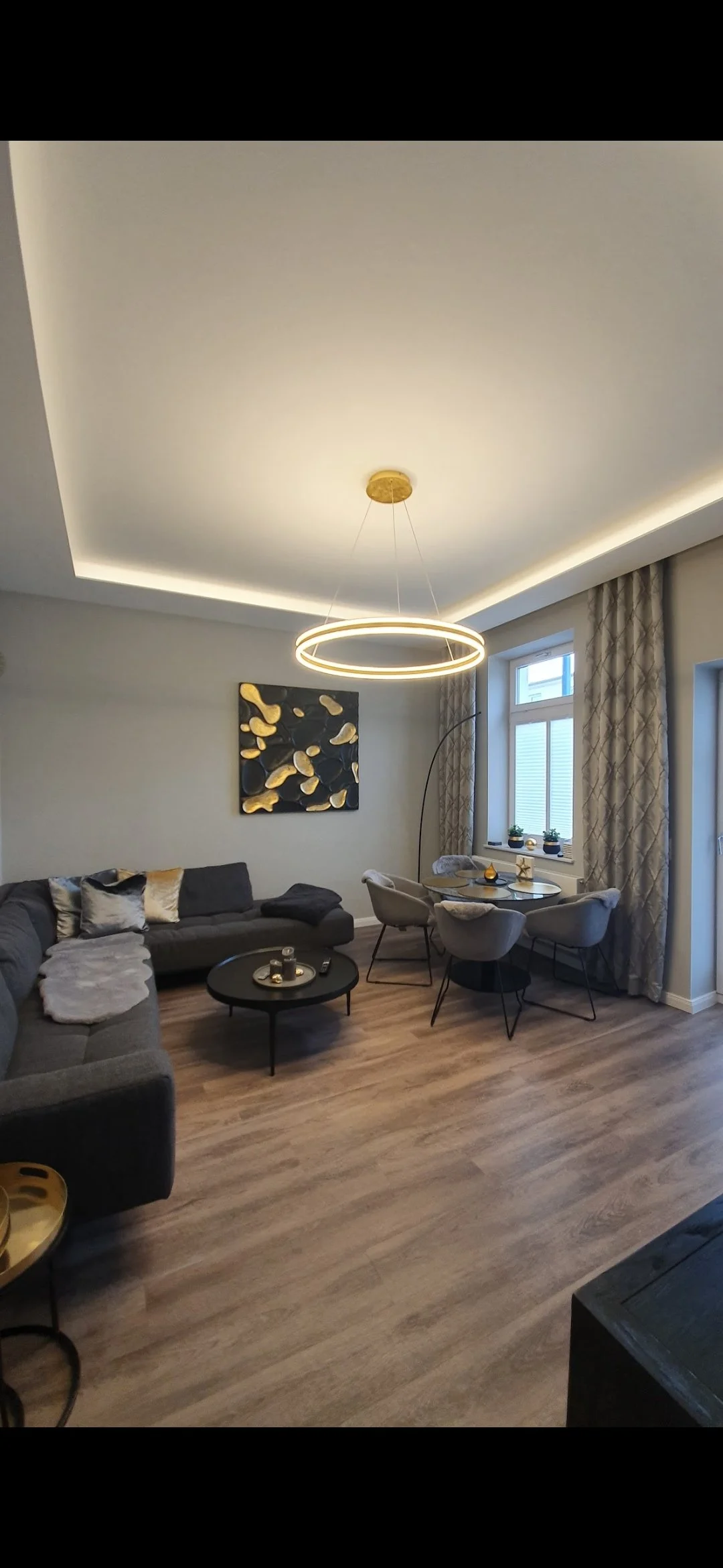 Modernes Wohnzimmer mit grauer Couch, runder schwarzen Couchtisch, Wandbild mit goldfarbenen Akzenten, Esstisch mit vier Stühlen, Fenster mit Vorhängen, dekorative Gegenstände auf der Fensterbank, hängende kreisförmige Hängeleuchte, Holzfußboden.