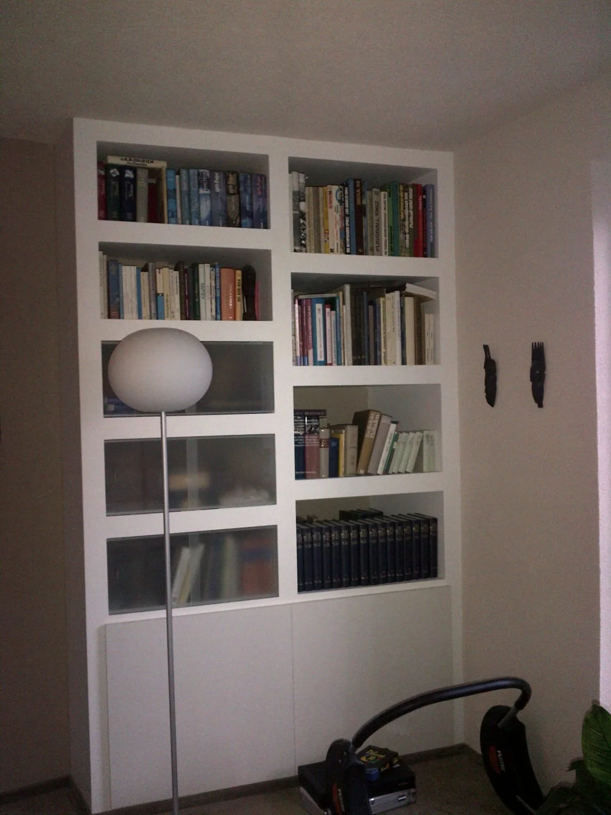 Ein weißer Bücherregal mit mehreren Fächern, gefüllt mit Büchern. Daneben steht ein moderner Stehlampe. In der Nähe befinden sich auf dem Boden ein Staubsauger und ein Werkzeugkasten, ein Stuhl ist im Vordergrund sichtbar.