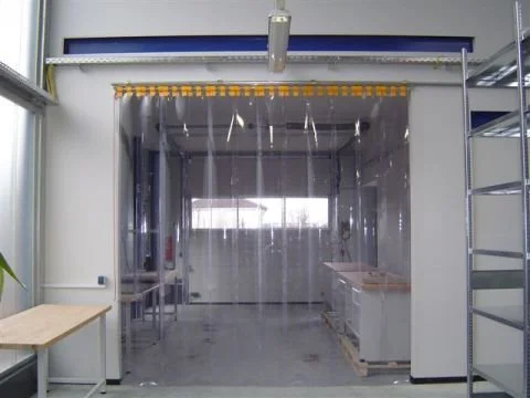 Eine transparente Abtrennung in einer Werkstatt oder Fabrik, mit einer blauen Vorrichtung an der Decke, gelben Vorhängen oben und einem Tisch auf der linken Seite.