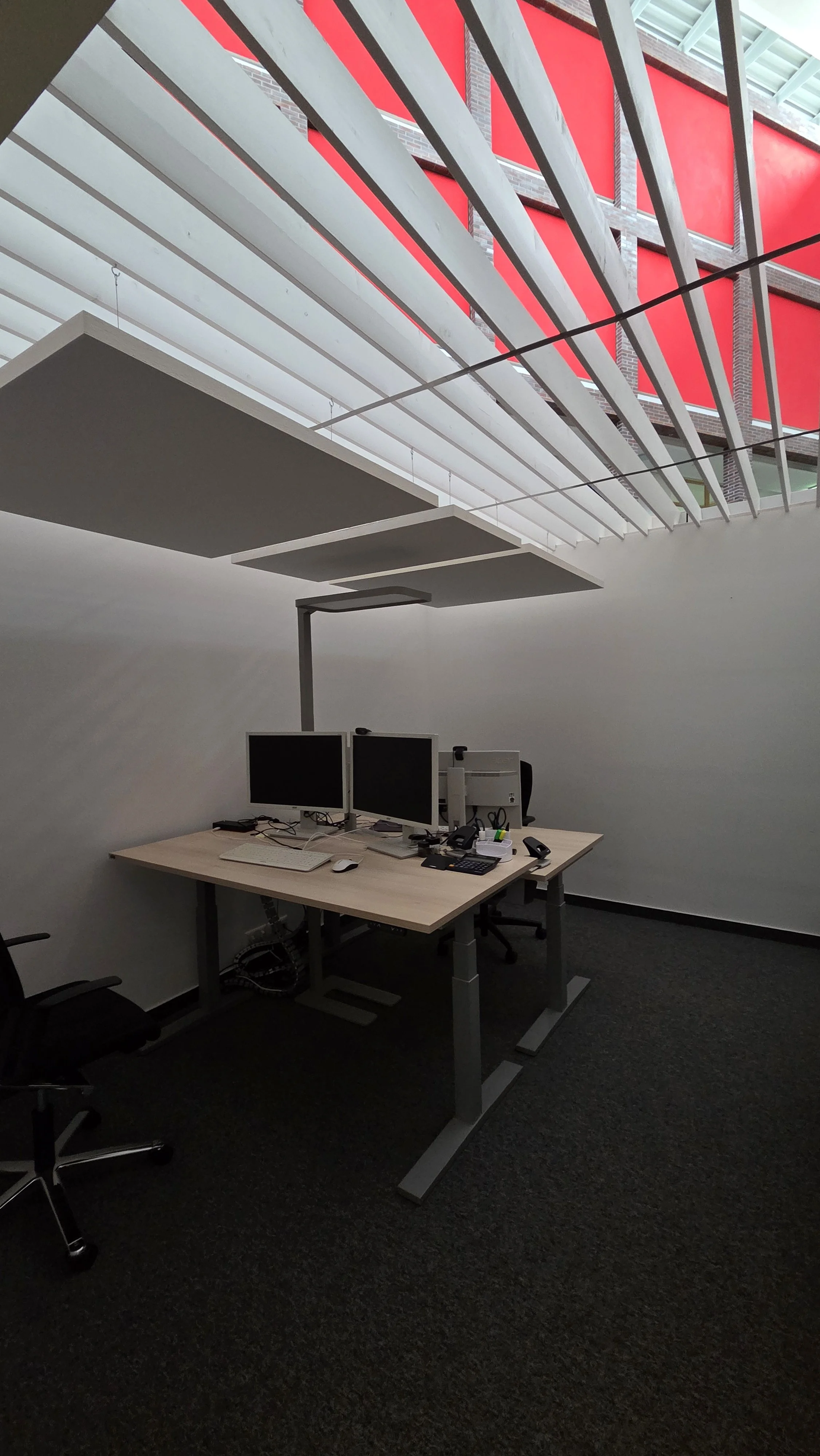 Ein Büro mit einem Schreibtisch, zwei Computerbildschirmen, einer Tastatur, einer Maus, Telefonen und Büromaterialien. Das Büro hat eine dunkle Teppichboden und weiße Wände, mit einem großen Fenster, das im oberen Bereich rote Fensterläden zeigt.